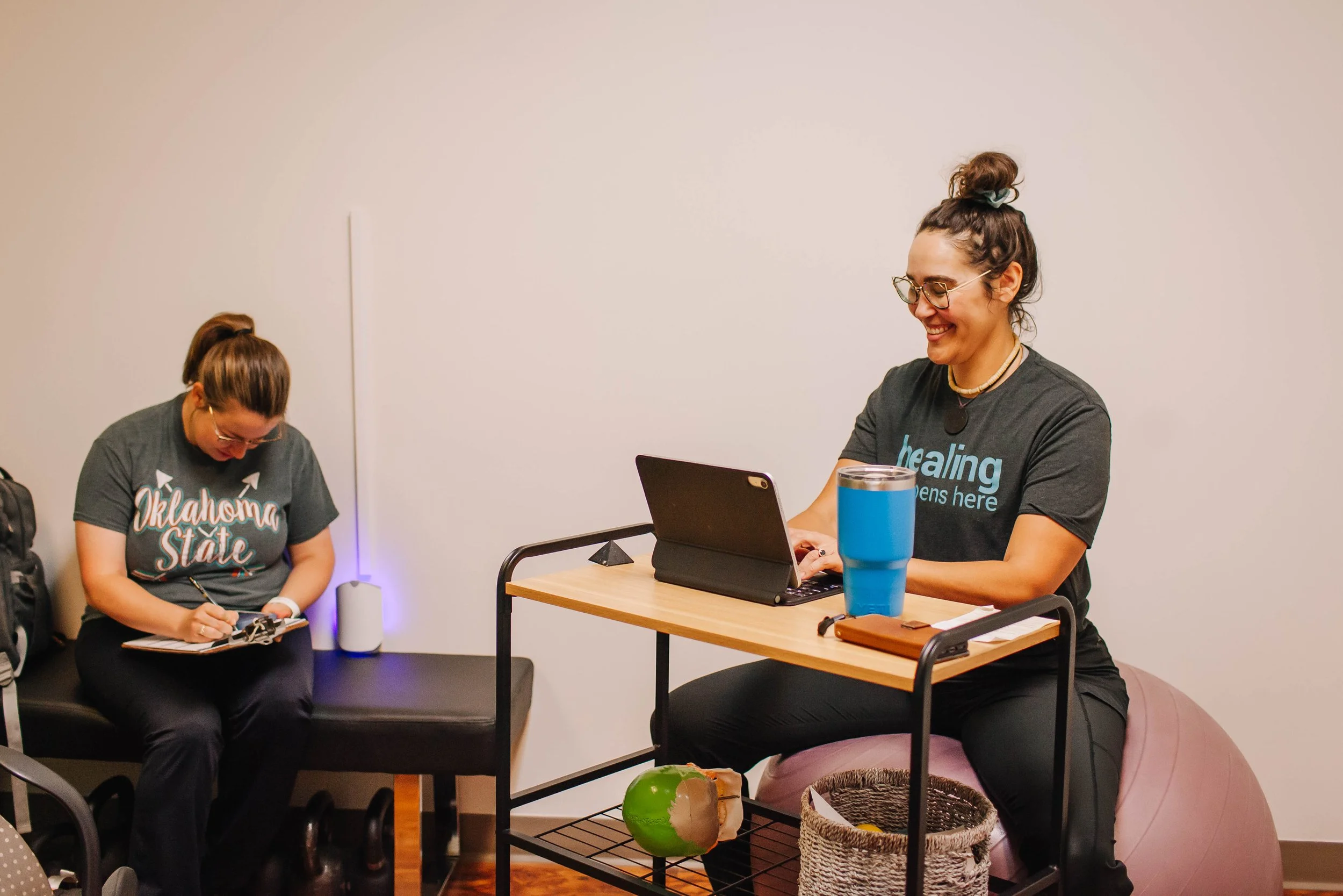 Two women sitting at a table in a room with plain white walls. The woman on the right is smiling, wearing glasses, a bun hairstyle, a necklace, and a dark t-shirt with blue text. The woman on the left is looking down at her phone, wearing glasses, a gray t-shirt with Oklahoma State written on it, and has her hair in a ponytail. There is a tablet on the table, a large blue water bottle, a brown wallet, a small bag, a half-eaten coconut, and a woven basket nearby.