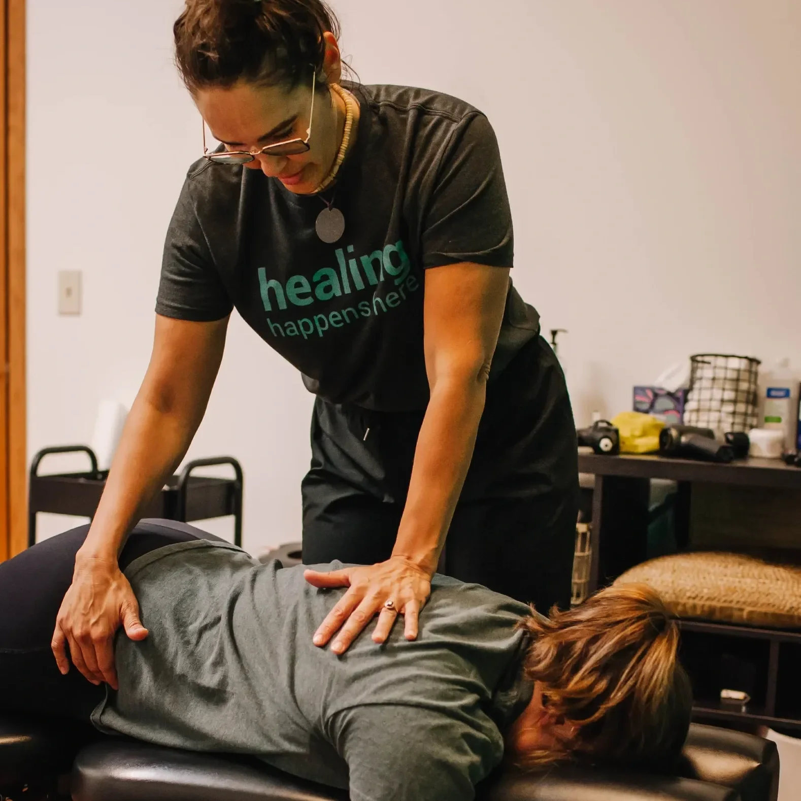 A woman providing a massage or chiropractic adjustment to another person lying face down on a table. The woman is wearing glasses and a t-shirt that says 'healing happens here.' She is using her hands to apply pressure to the person's back.