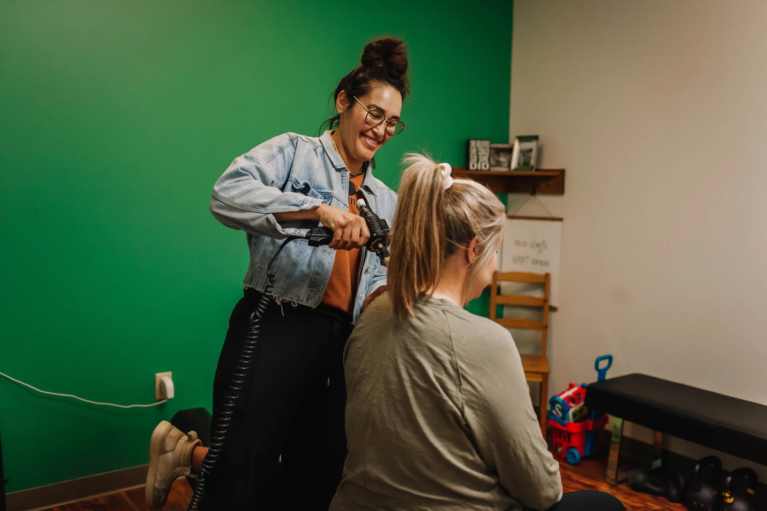 A woman is styling a girl's hair using a curling iron in a room with a green wall and wooden flooring. The woman is smiling and wearing glasses and a denim jacket.