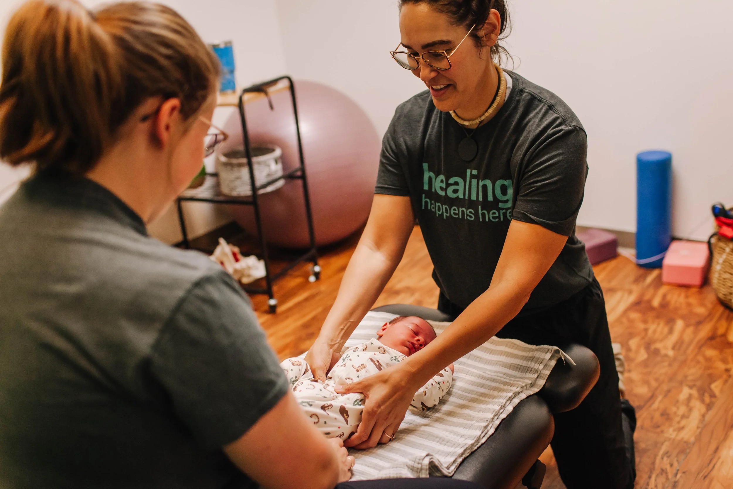 A woman with glasses and a necklace is leaning over a baby lying on a padded surface, performing a routine check or activity while another person watches. The woman is wearing a black T-shirt that says 'healing happens here'.