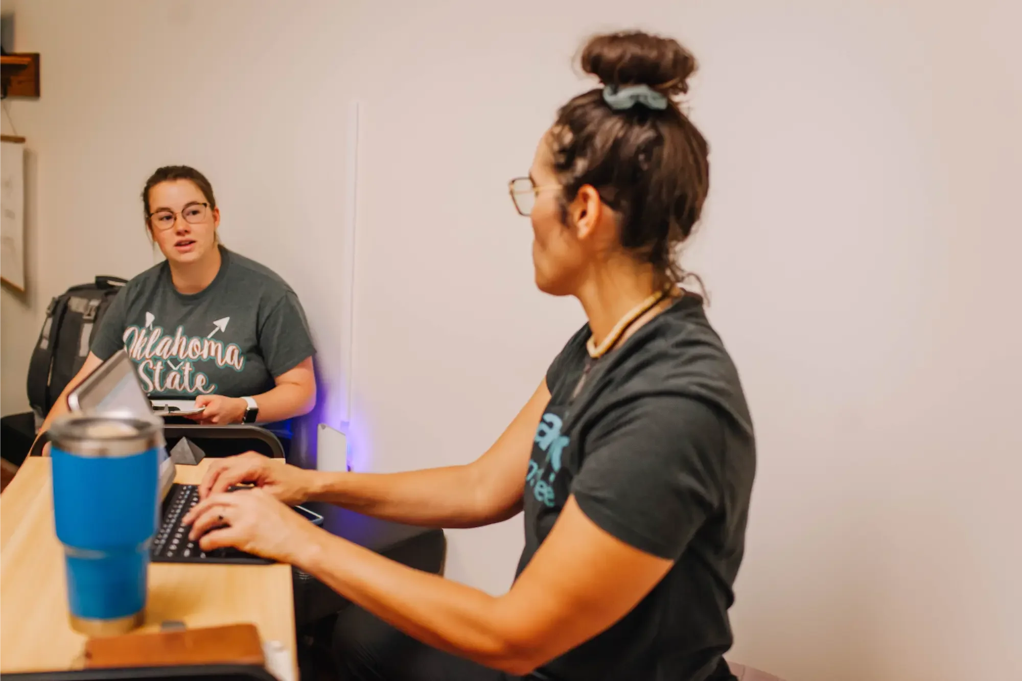 Two women sitting at a table working on laptops in a room with plain white walls; one is wearing glasses, a dark T-shirt, and has her hair in a bun with a scrunchie, while the other is wearing a University of Oklahoma T-shirt and glasses.