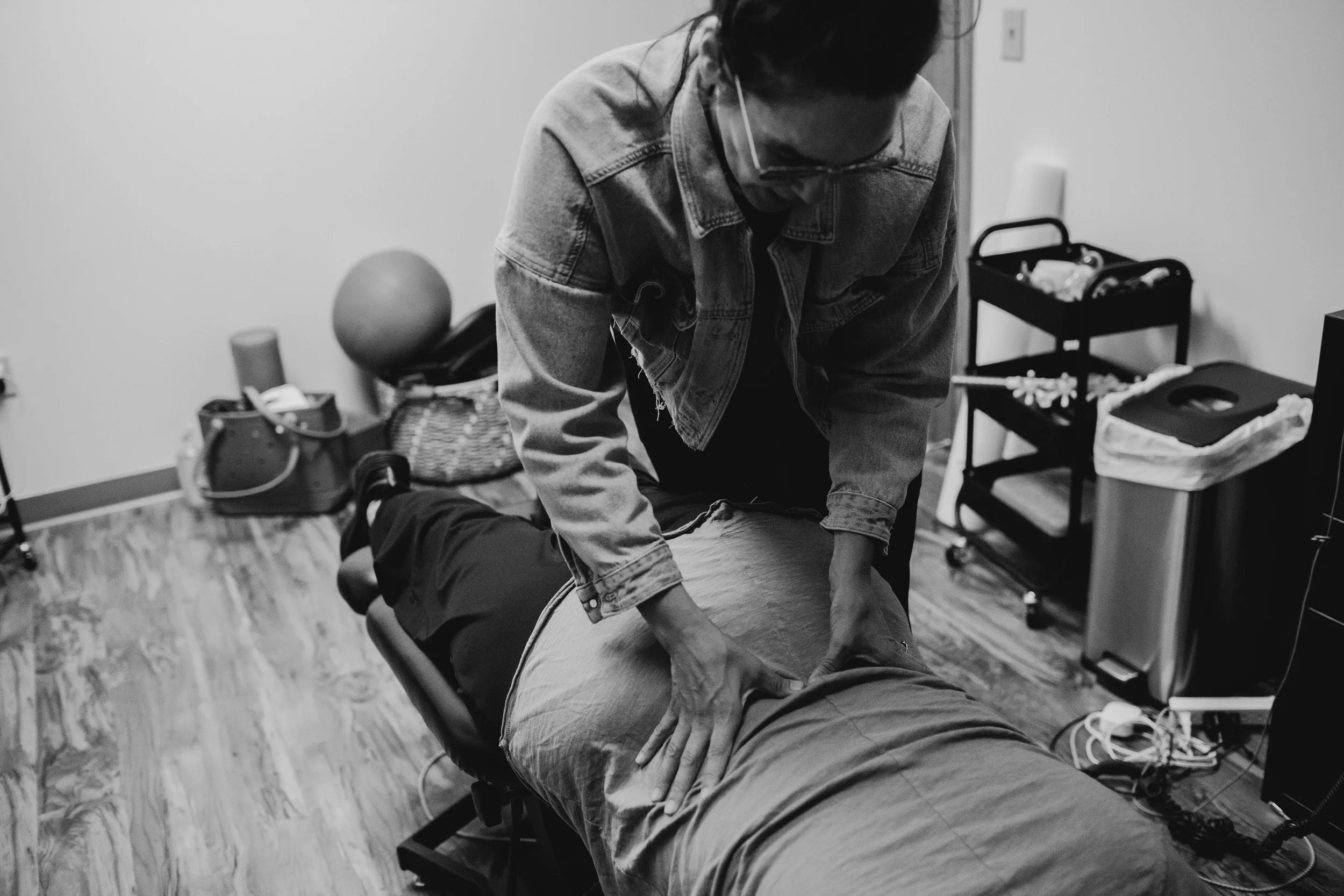 Black and white photo of a person giving a massage to a client lying face down on a massage table in a room with medical or therapy equipment.