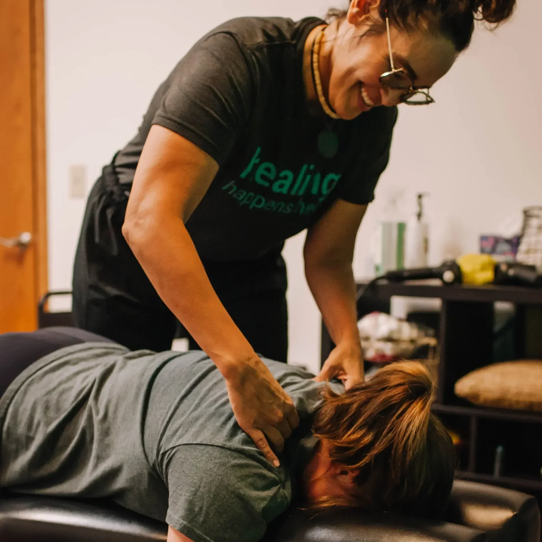 A woman receiving a massage from a therapist in a massage room, with the therapist leaning over and applying pressure to the woman's back.