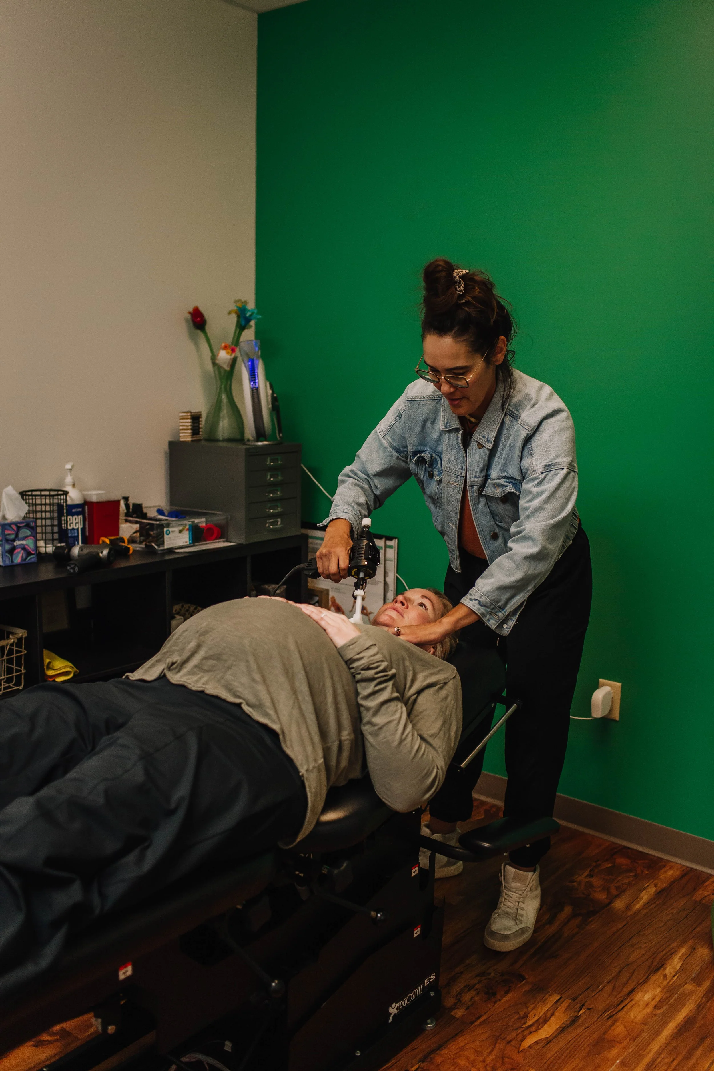 A woman receiving a treatment or therapy from a professional in a room with a green accent wall. The woman is lying on a massage or therapy table, and the professional is using a handheld device near her face. The room has various supplies and decorative items on shelves and a counter in the background.