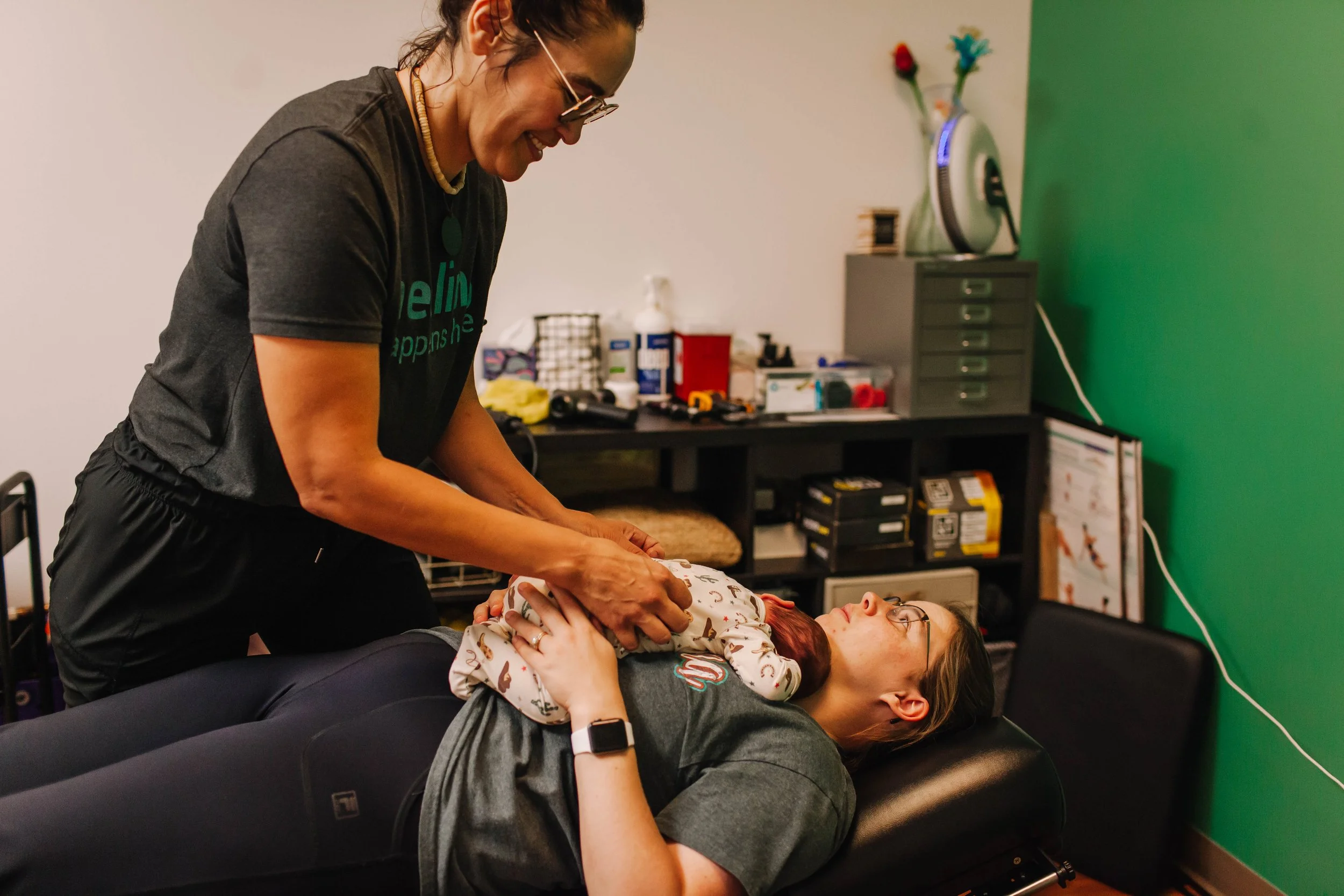 A woman performs a medical or chiropractic adjustment on person lying on a table, with a young girl lying on her back on the woman's chest, in a room with a green wall and various medical supplies in the background.