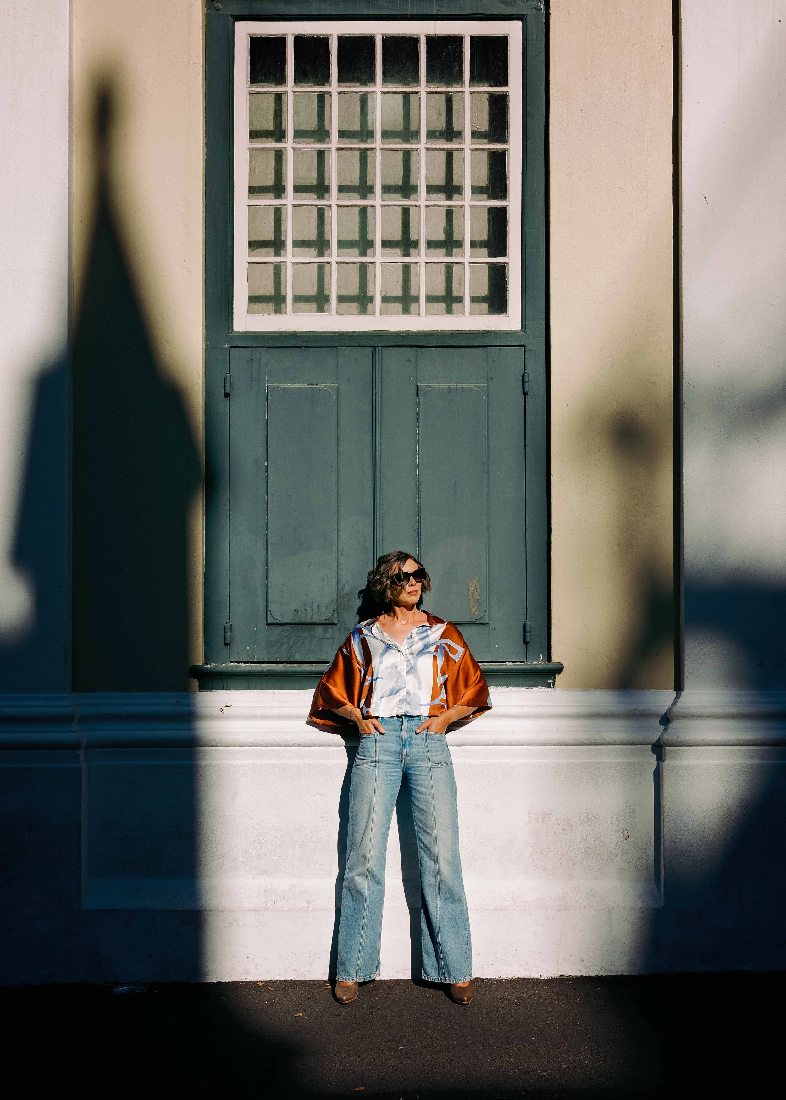 Jessica R., a woman with curly hair wearing sunglasses, a colorful patterned shirt, and jeans, standing outdoors against a white wall and a green door, with shadows cast on the wall.