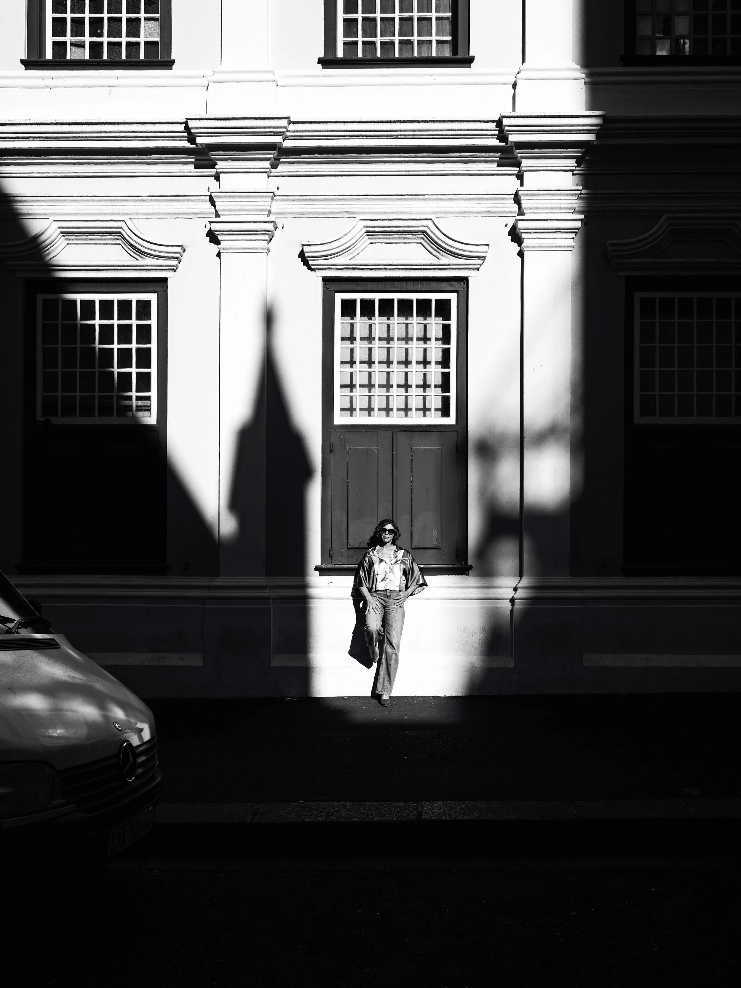 Black and white photo of Jessica R.,  a woman with sunglasses, standing with hands on hips in front of an ornate building with large windows and decorative architectural details, partially illuminated by sunlight.