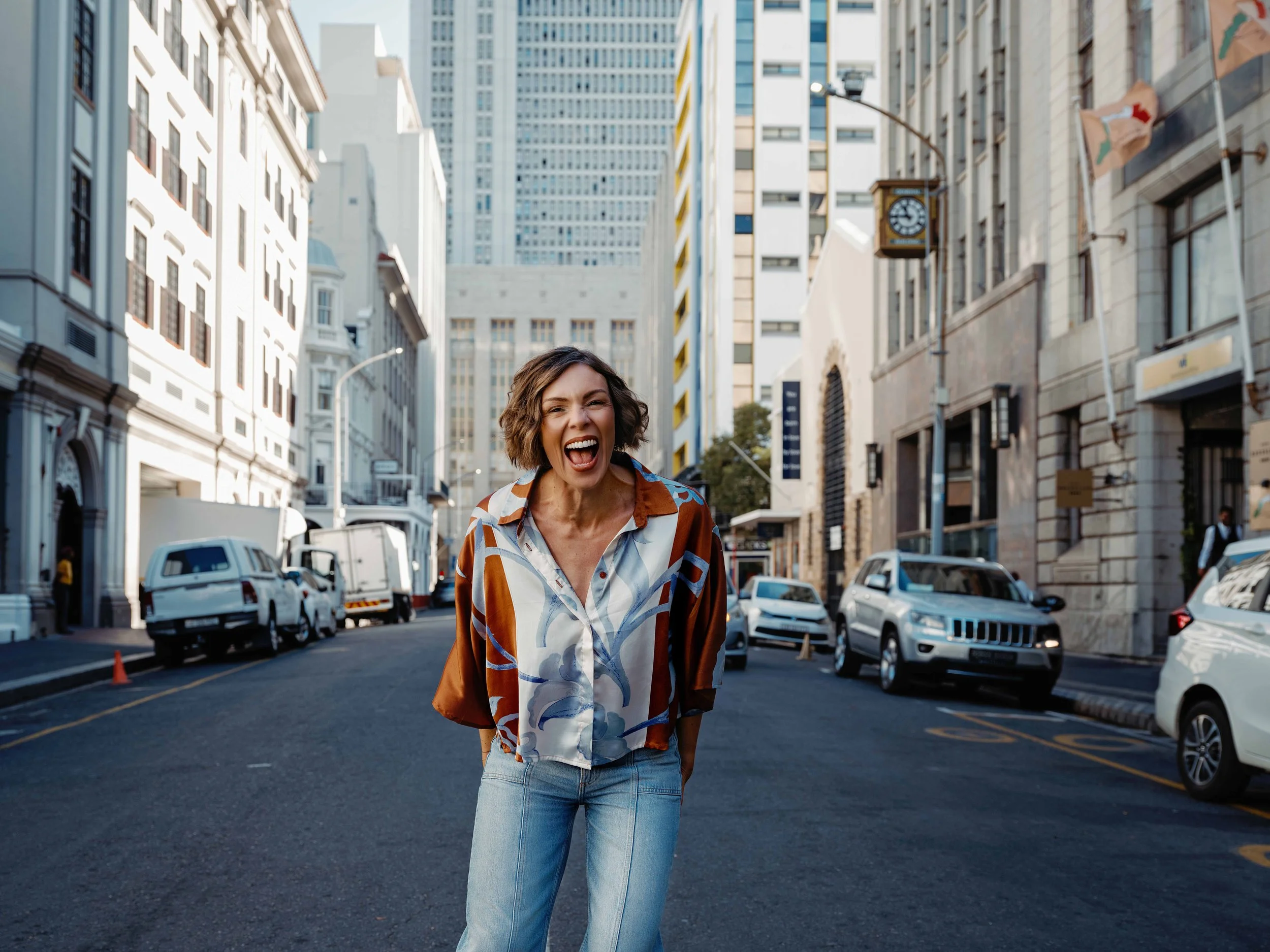 A woman with short curly hair smiling and walking down a city street surrounded by tall buildings, parked cars, and flags.