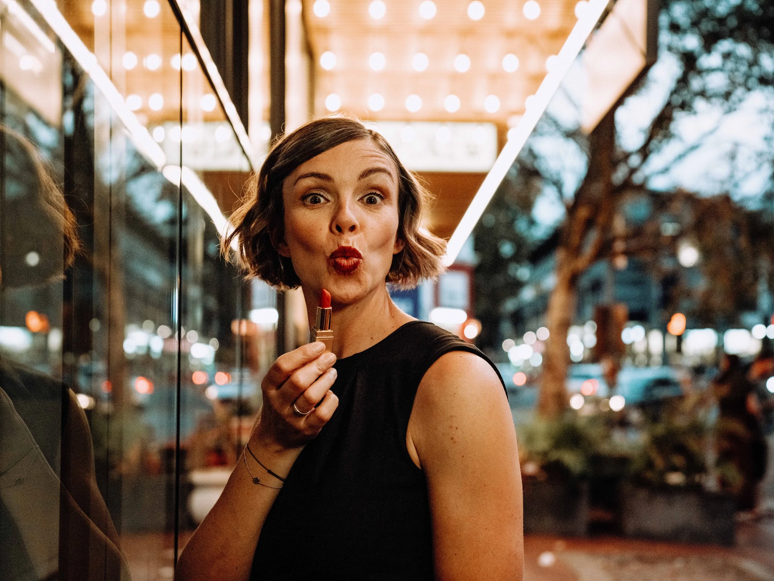 Jessica R., a woman with short brown hair, red lipstick, and a black top holds a lipstick tube near her lips. She is standing outside or near a reflective glass surface at night, with blurred lights and trees in the background.