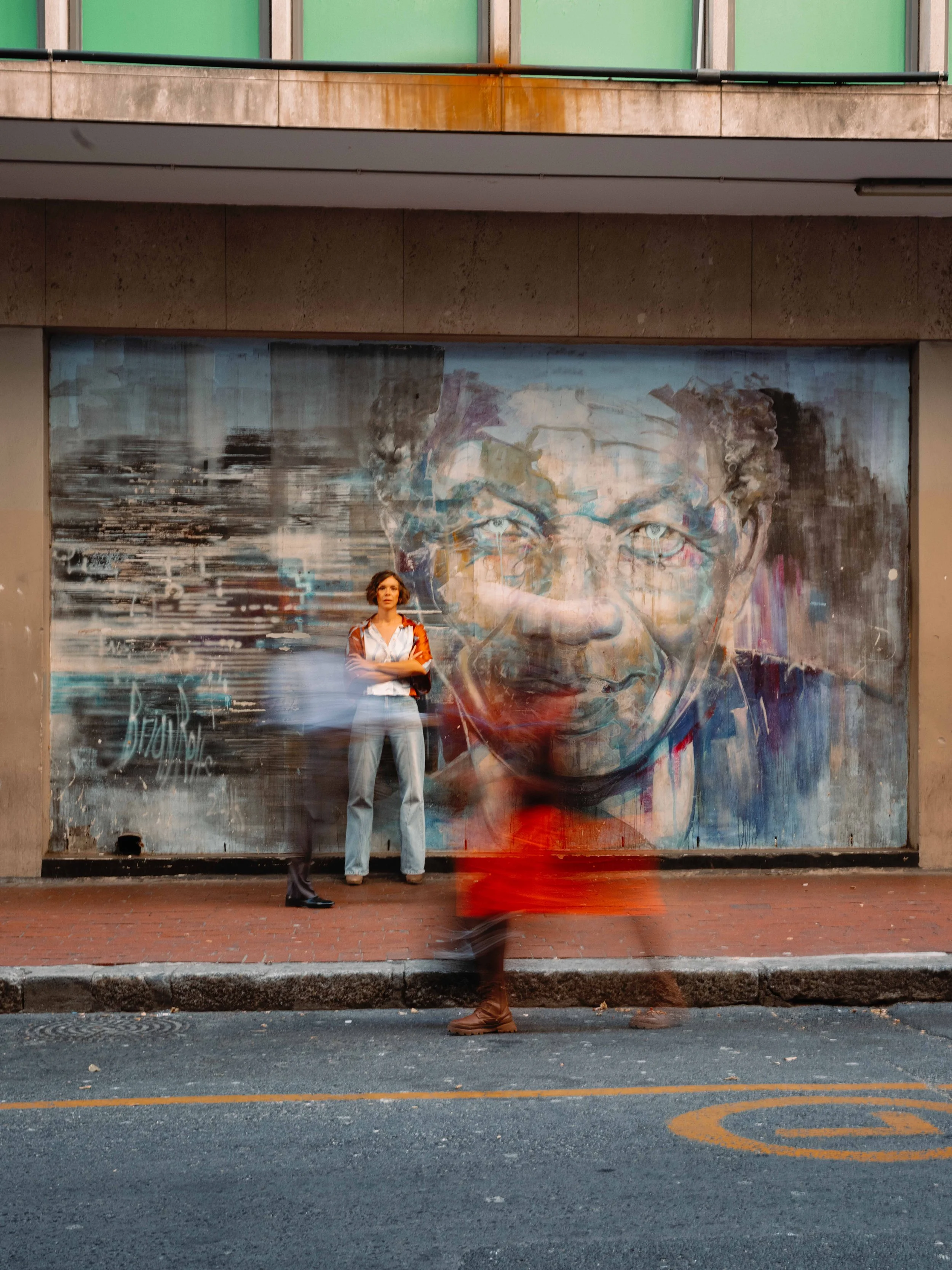 Jessica R., a woman standing in front of a large urban mural of a person's face, with a blurred person walking by in front of her. The scene is on a city sidewalk with a brick section and street markings visible at the bottom.