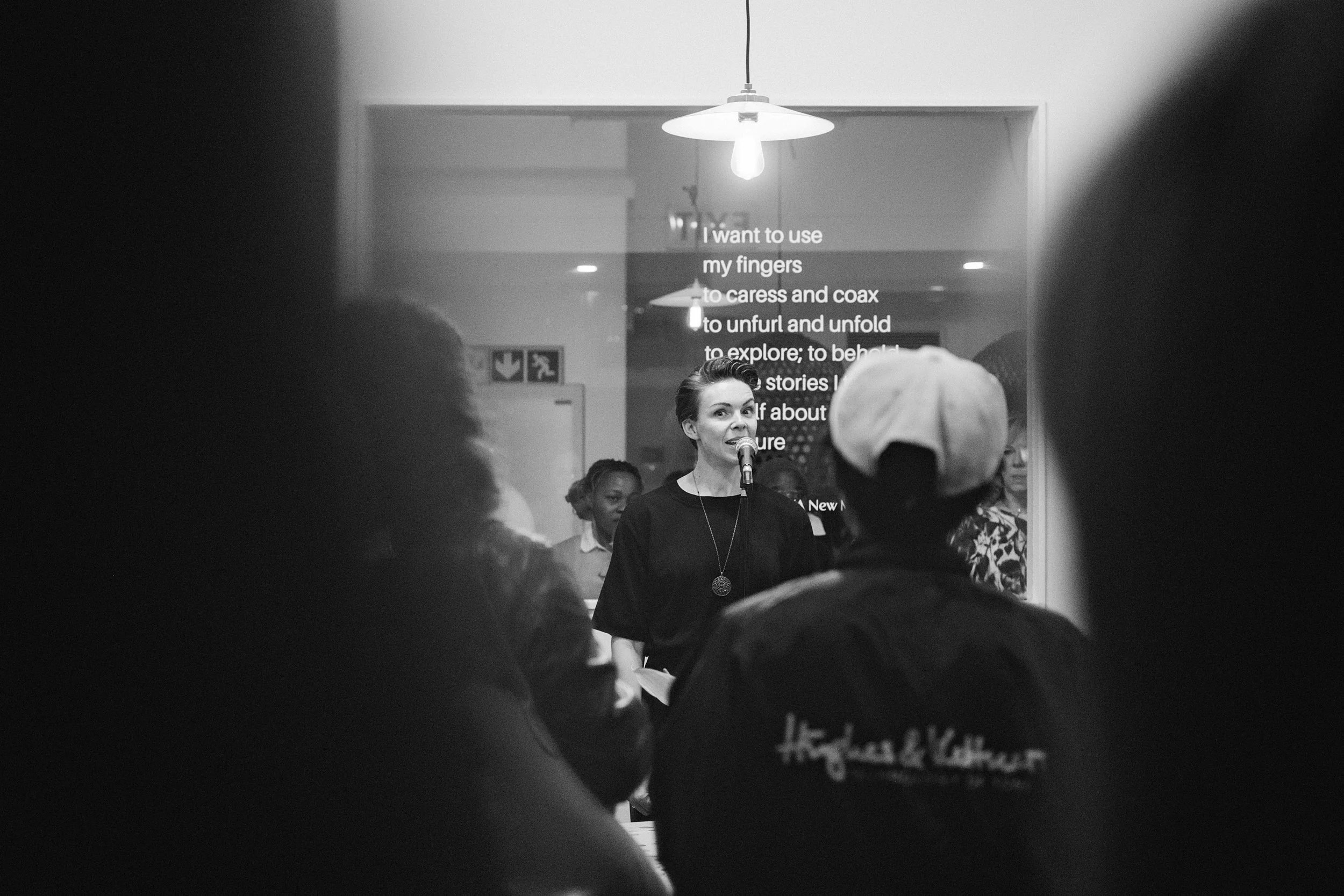Jessica R., a woman is speaking into a microphone during an indoor event, with a group of people listening. There is a hanging light fixture above and a display with text behind her, partially visible.