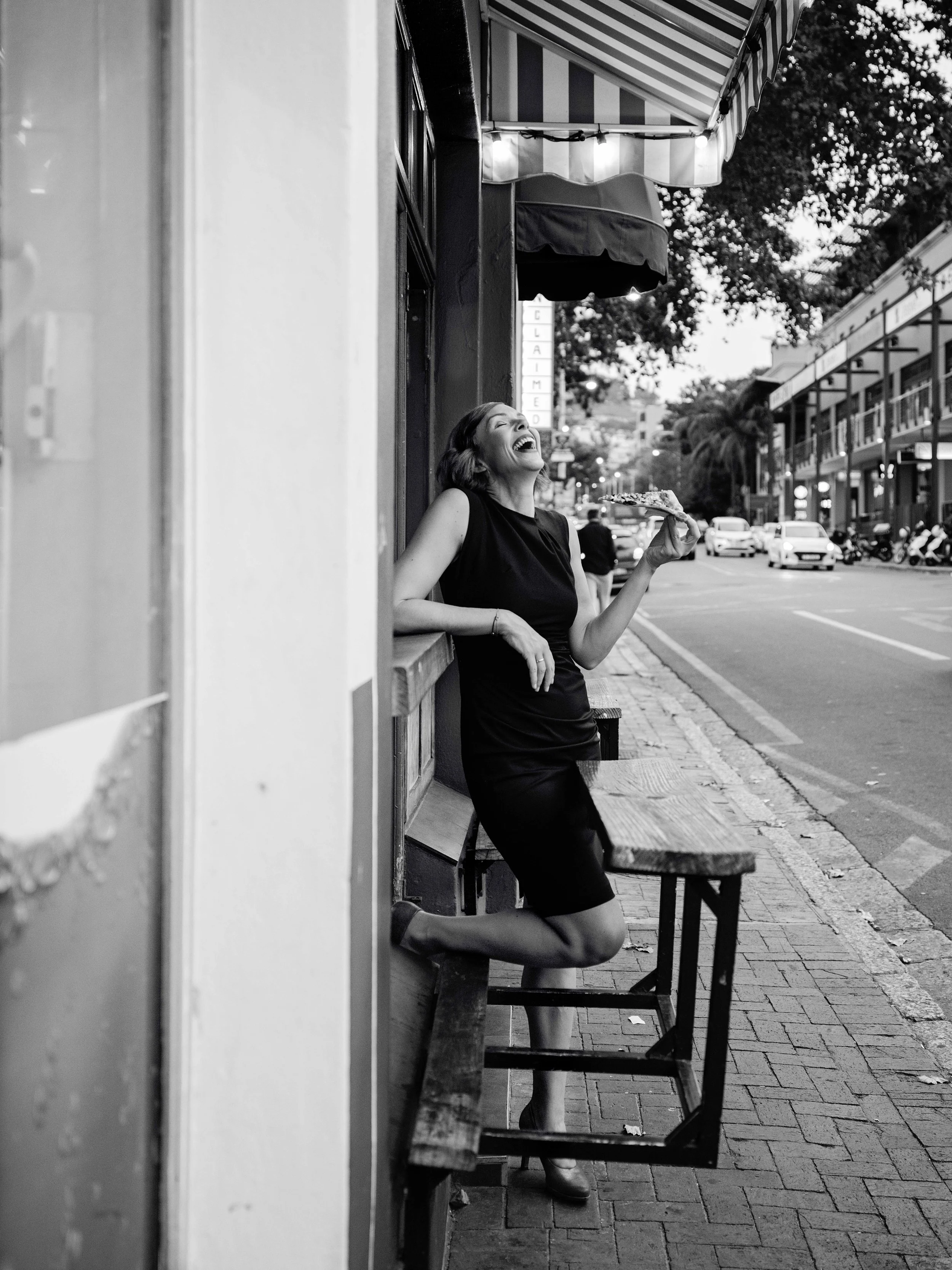 Jessica R., a woman standing outside a storefront, laughing and holding a slice of pizza in her right hand, with the street and parked cars visible in the background. The image is black and white.