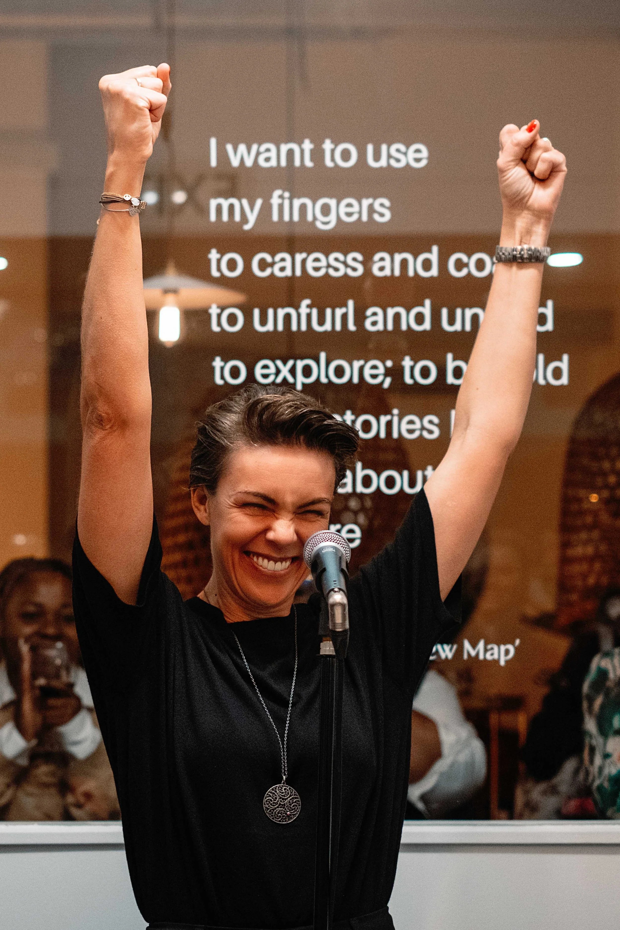 Jessica R., a woman smiling and raising her arms in celebration while speaking into a microphone at an indoor event, with a screen displaying text behind her.