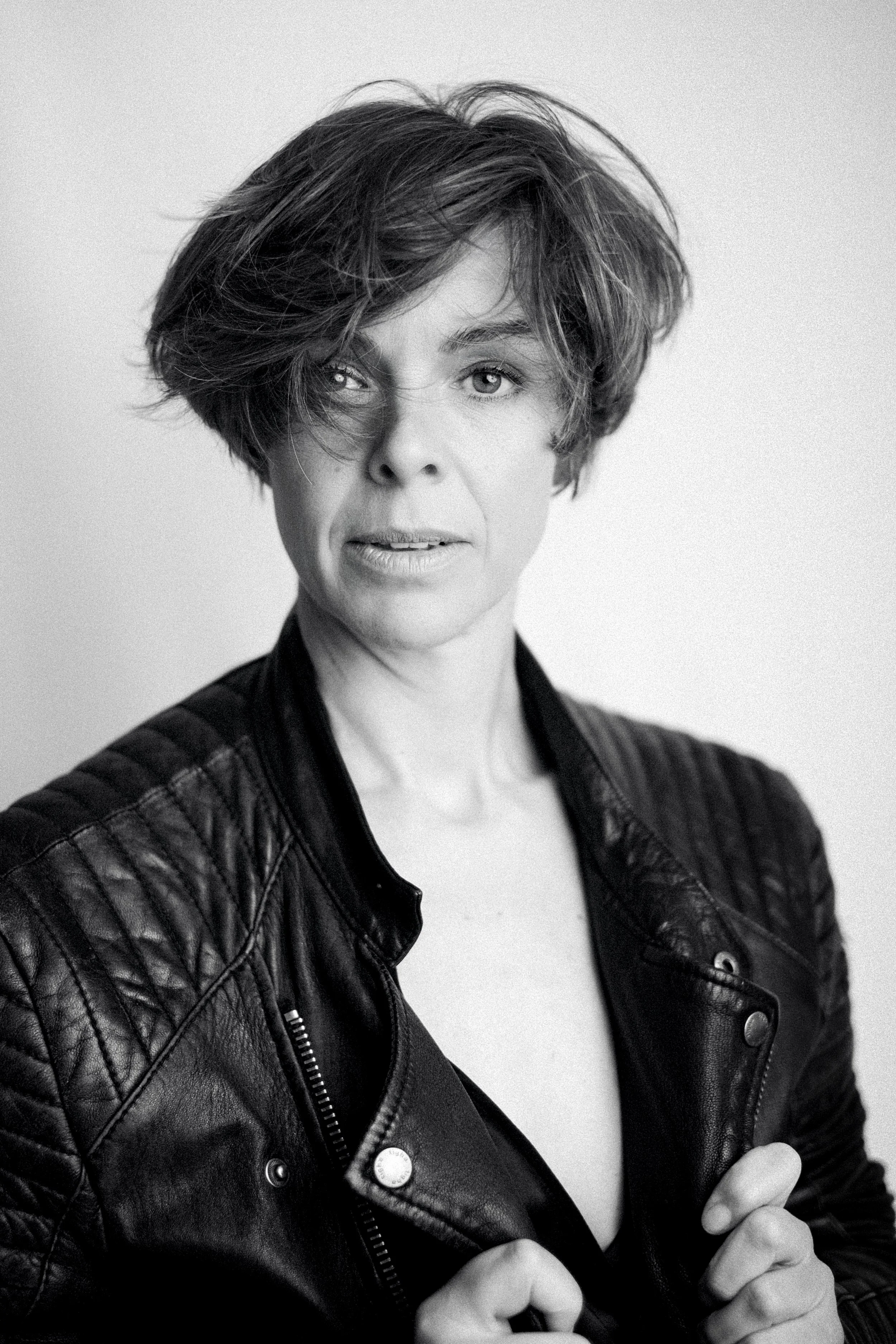 Jessica R., a woman with short, curly hair and light-colored eyes wearing a black leather jacket and a white top, posing with her fists extended in a confident stance.