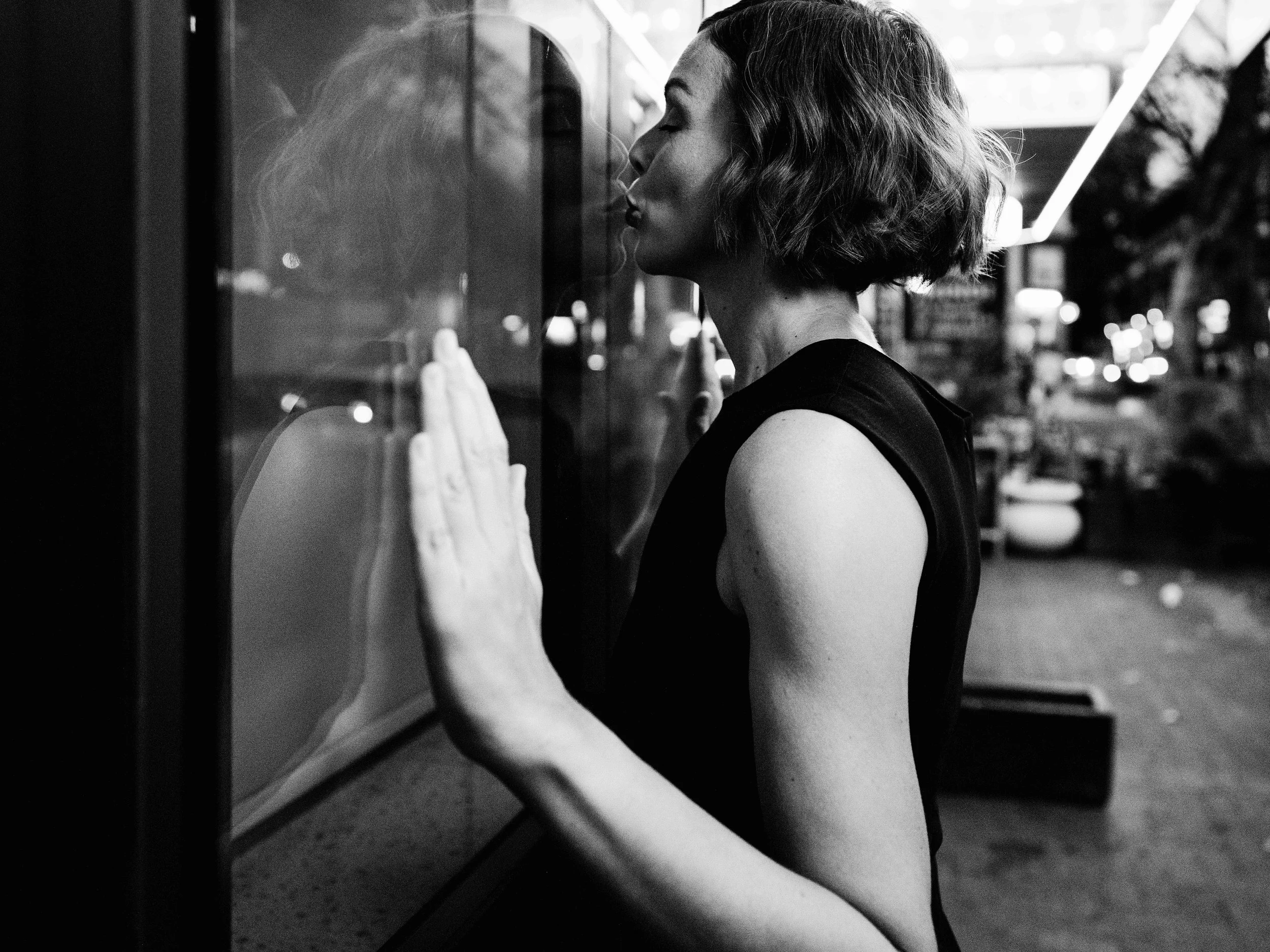 Jessica R., a woman with short hair in a black dress with sleeveless design, standing close to a vending machine, with her eyes closed, touching the machine with her hand, in an urban setting at night.