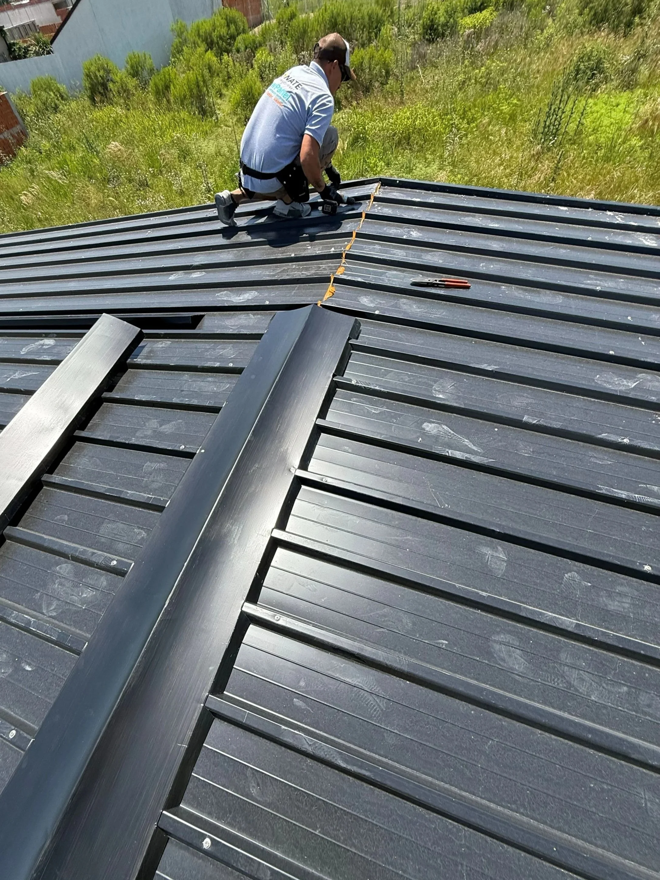 A man working on a metal roof, kneeling and measuring the roofing panels with a yellow tape measure, tools nearby on the roof, in a grassy area with sparse buildings in the background.