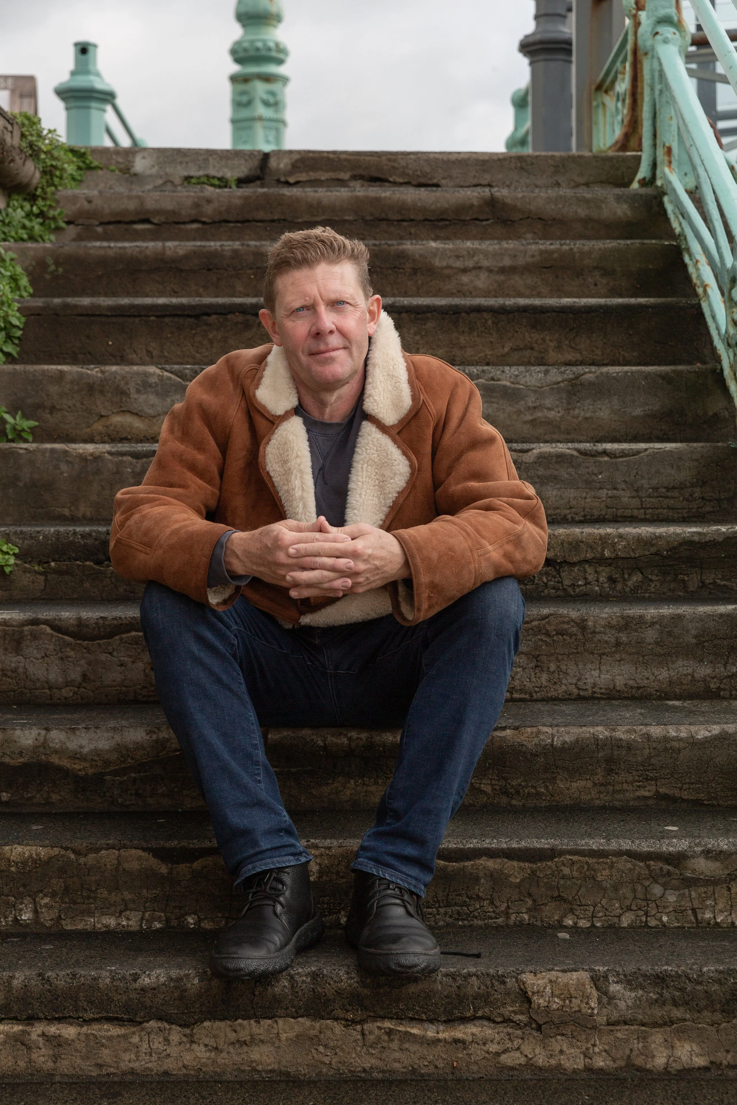 Peter Hanington, author, sitting on  stone stairs in Brighton England, wearing a brown jacket with a fur collar, blue jeans, and black boots. He has short hair and is looking at the camera with his hands clasped.
