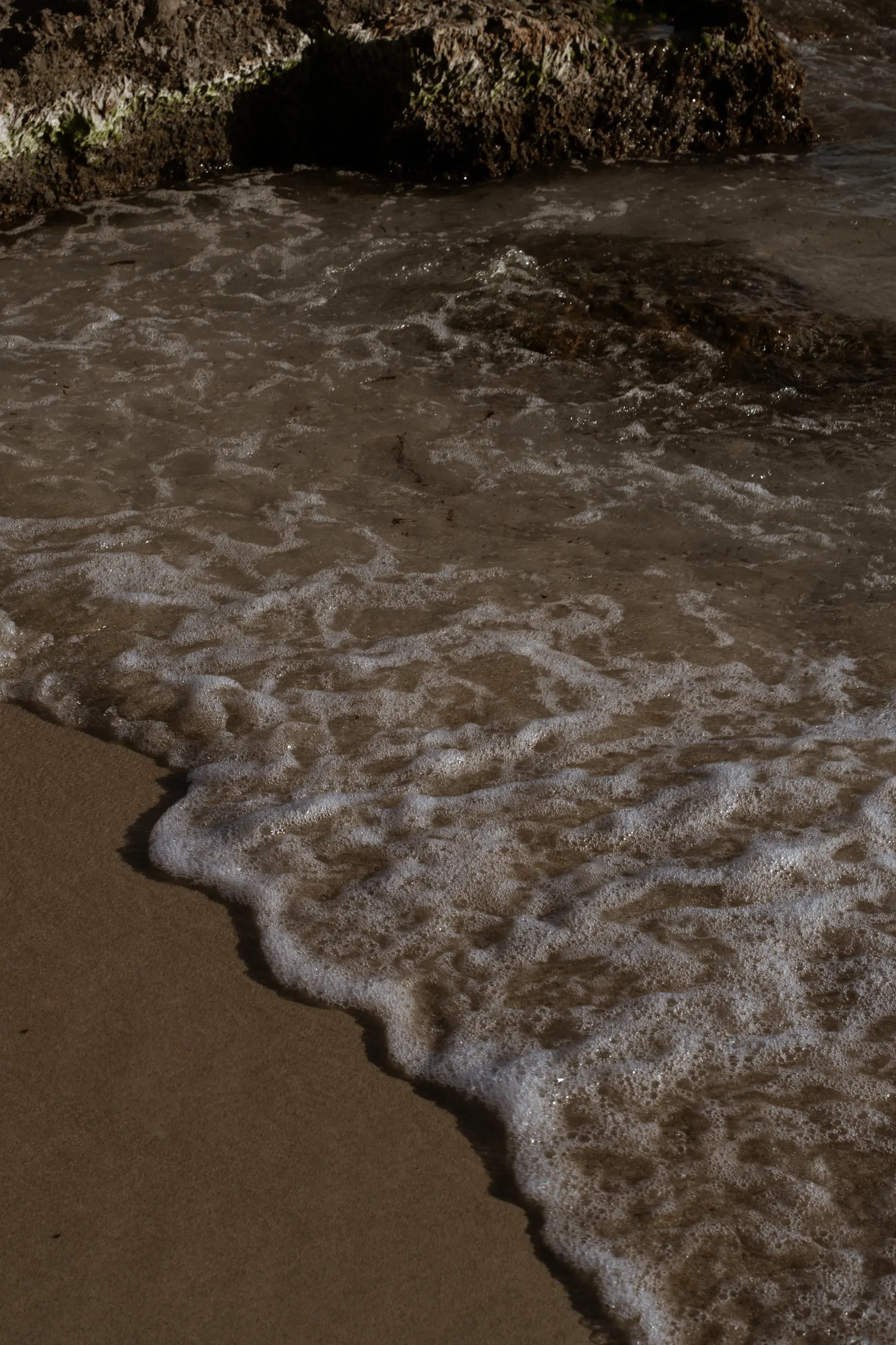 Close-up of waves washing onto a sandy beach with rocks in the background.