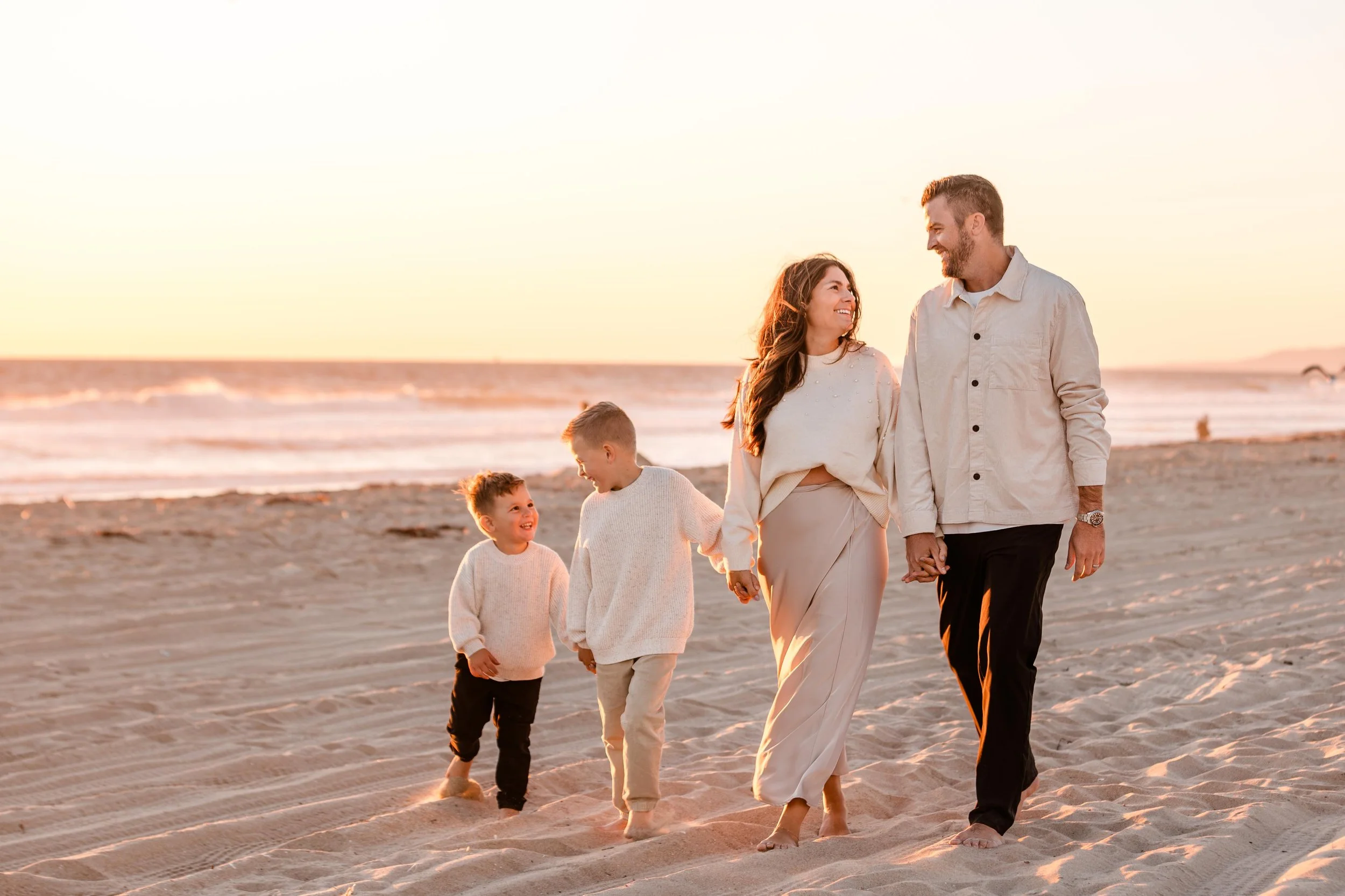 A family of four walking along the beach at sunset, holding hands and smiling at each other.