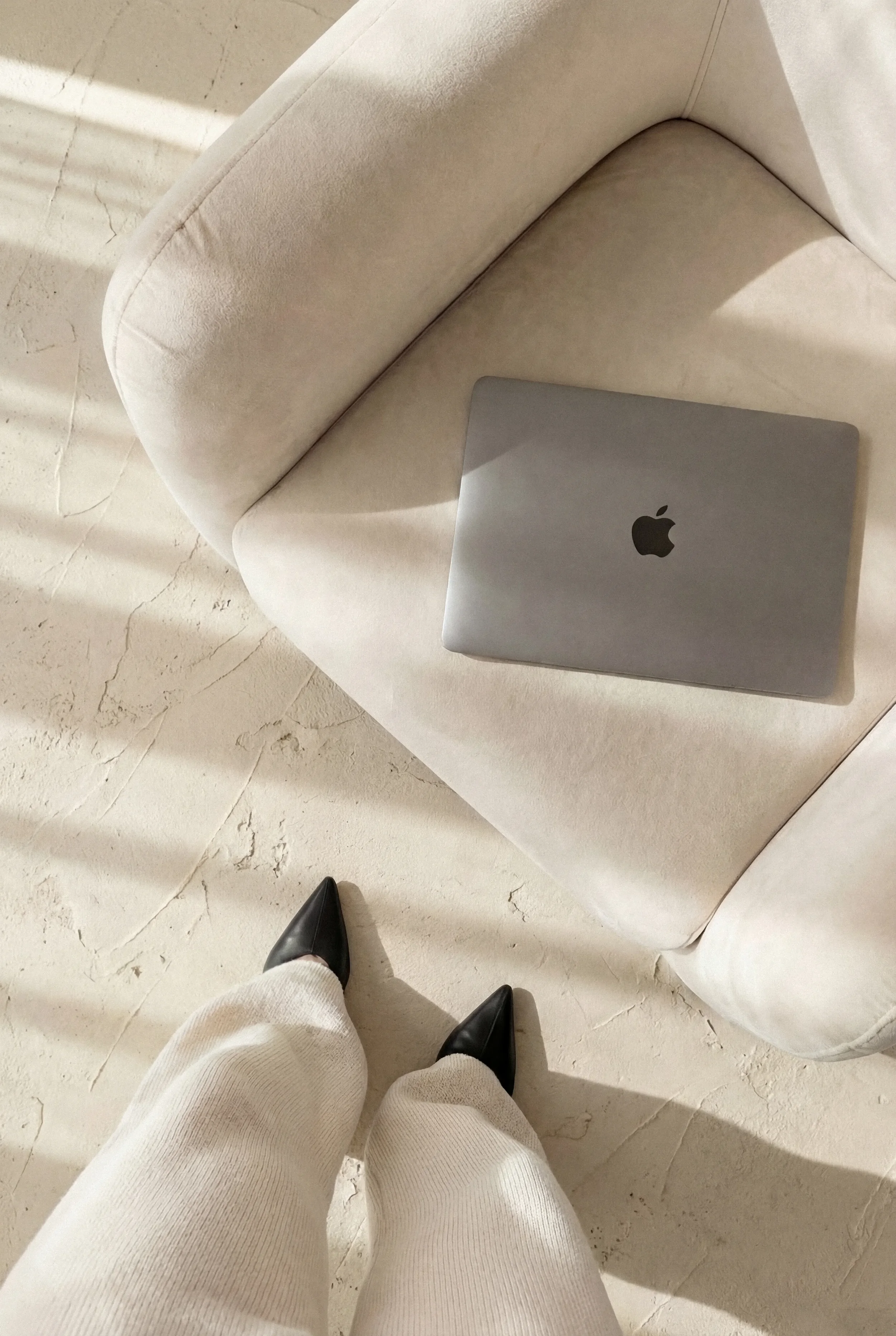 A person wearing white pants and black shoes standing on a white textured floor. A grey MacBook is on a white surface nearby.
