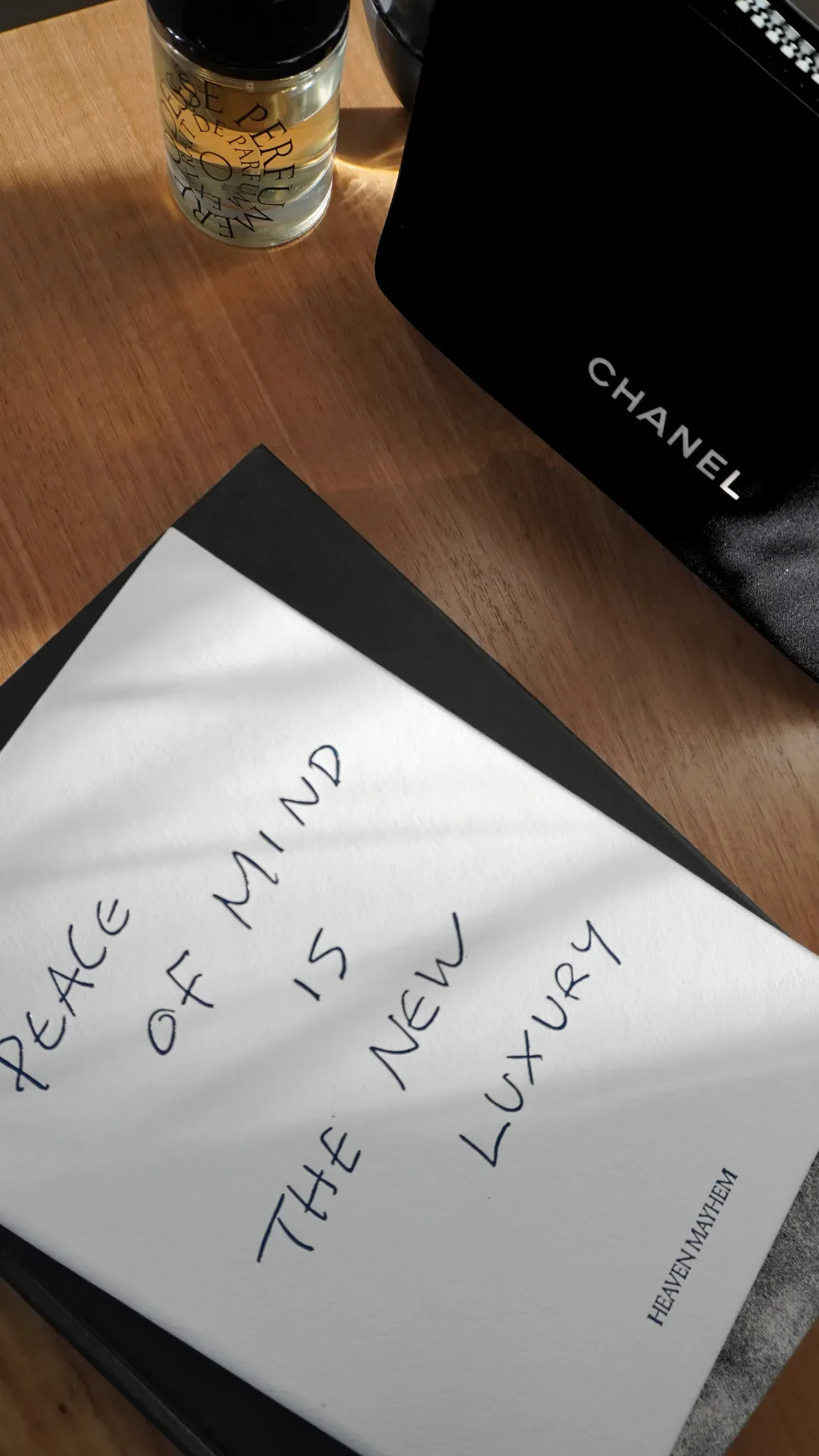 A handwritten note on white paper with the words "HEAVEN MAY ONLY BE THE AFTER 15 NOV 5, LUXURY". To the top left, part of a black-covered notebook or book is visible. On the top left corner, there is a container with a perforated label. To the top right, part of a black Chanel box is seen.