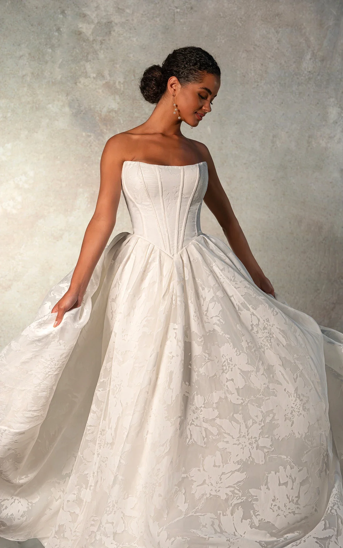 A woman wearing a strapless white wedding gown with a full skirt, standing against a textured light-colored background, with her head slightly tilted and eyes closed.
