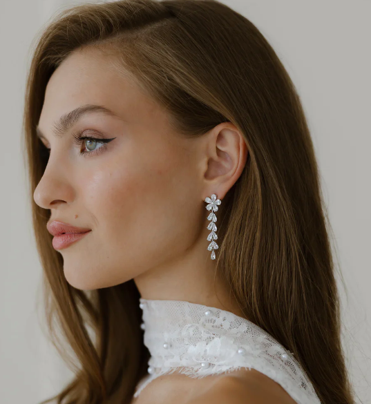 A woman with long brown hair wearing silver earrings and a white lace dress.