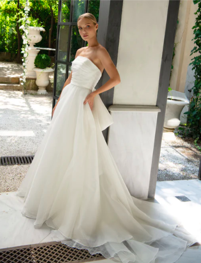 A young woman in a strapless white wedding gown standing outdoors on a bright day, with a modern architectural background and greenery.