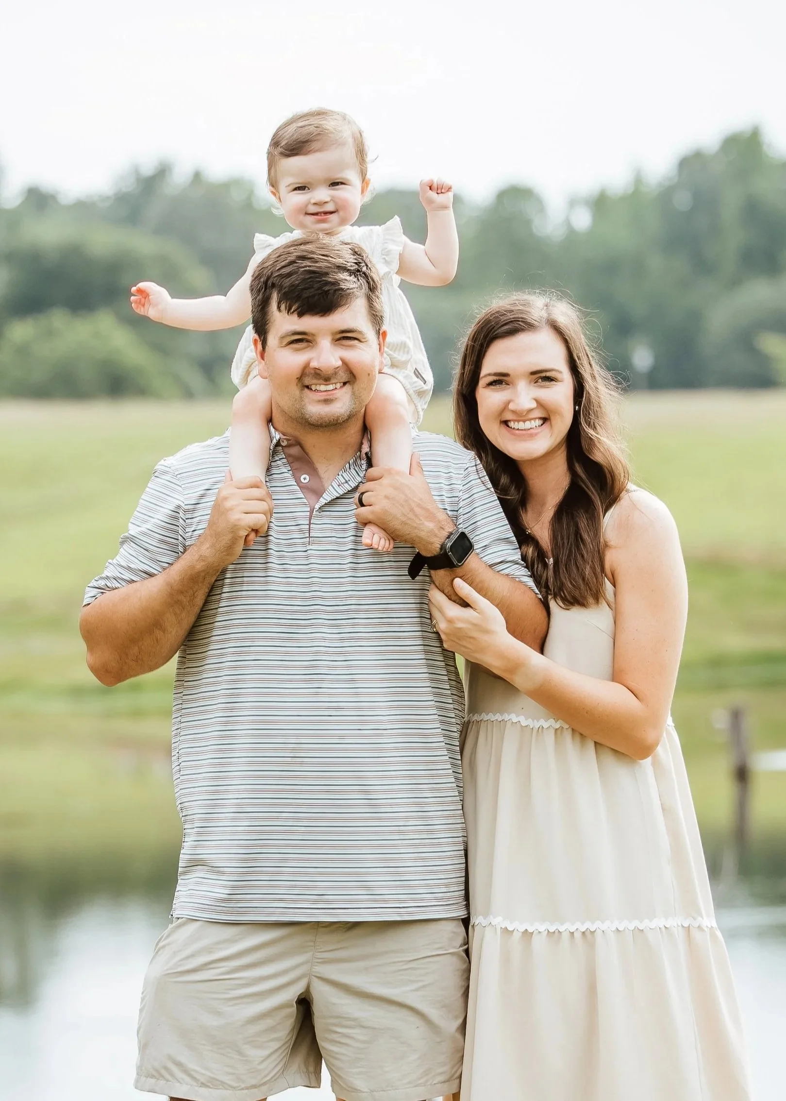 A happy family of three outdoors, with a father carrying a young girl on his shoulders and a smiling mother standing beside them, in a grassy field with trees in the background.