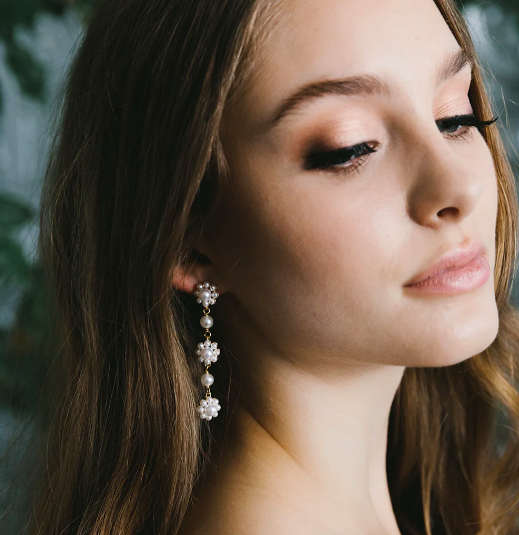 Close-up of a young woman with long brown hair, wearing a dangling pearl and floral earrings, with closed eyes and a slight smile.
