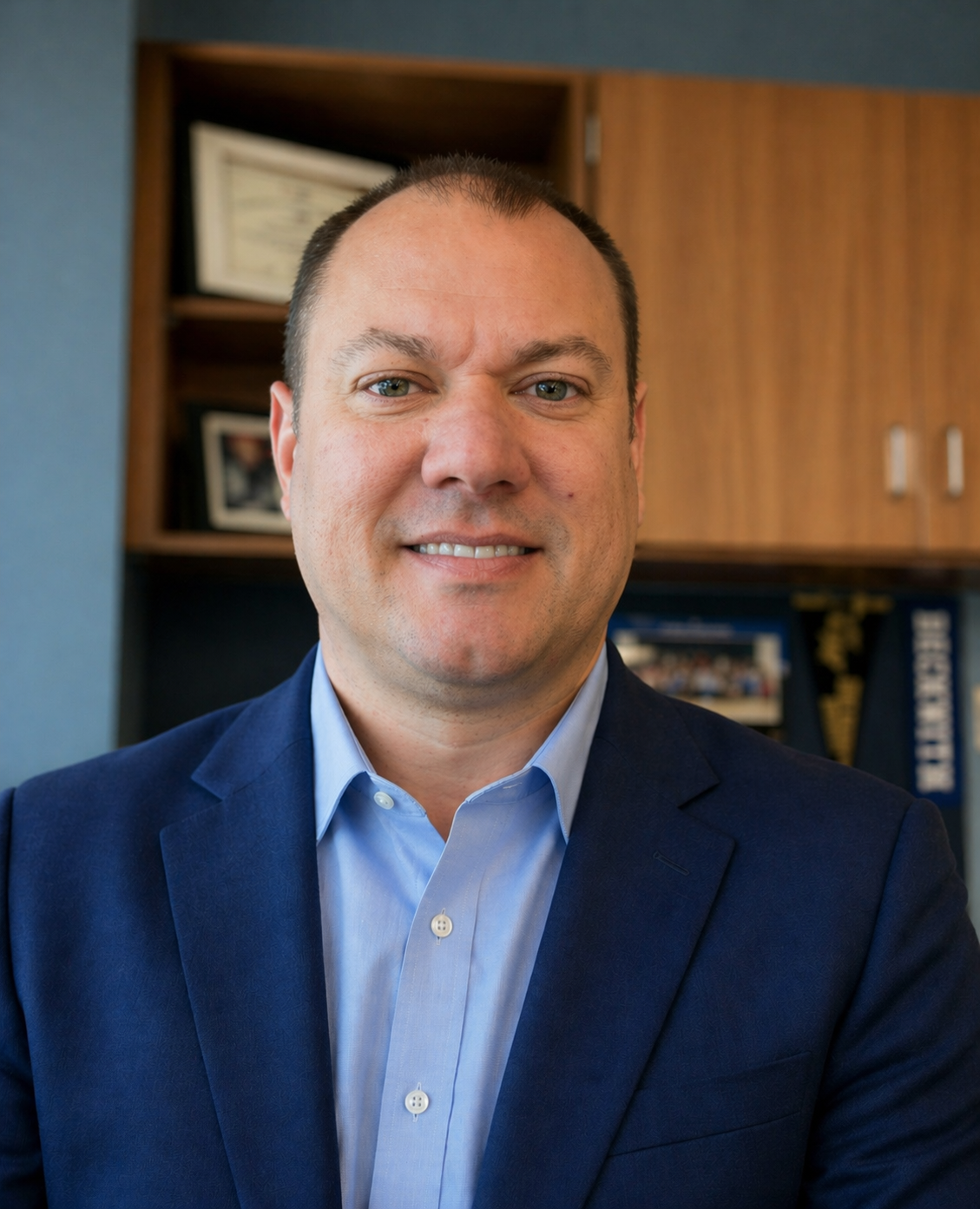 A professional man smiling, wearing a navy blue suit and light blue shirt, seated in an office with wooden cabinets and framed documents or pictures in the background.