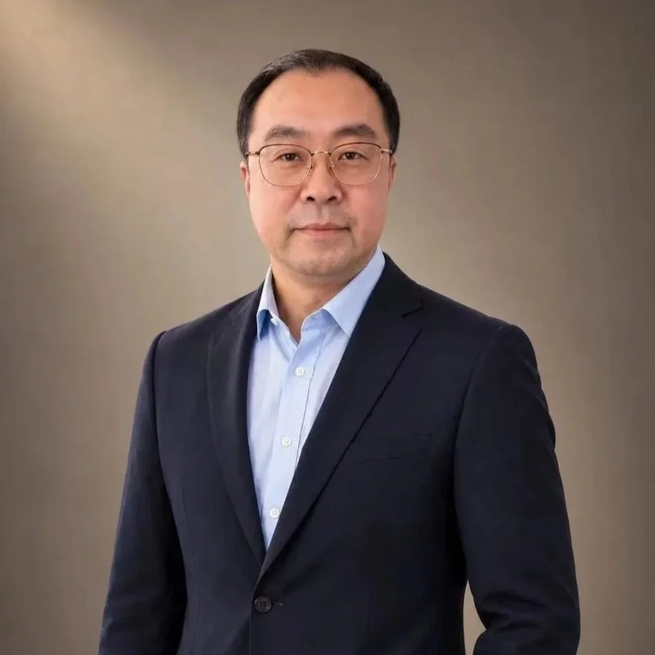 A man wearing glasses in a dark suit and light blue shirt stands against a plain background.