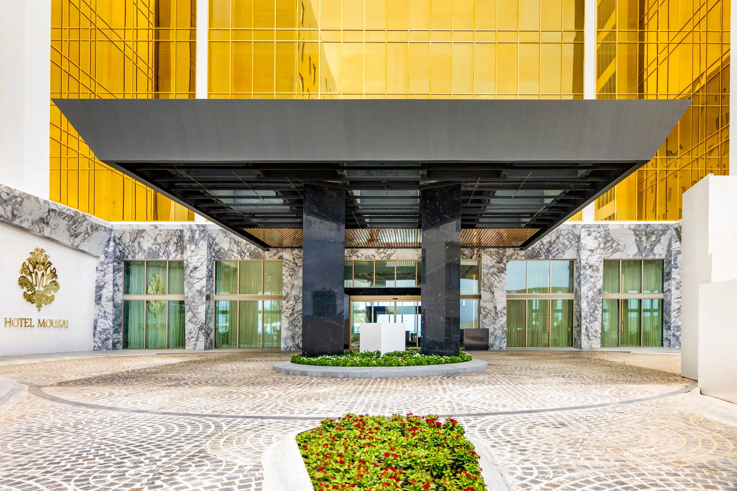 Exterior view of Hotel Mousai with a modern entrance, black marble pillars, green plants, and gold accents.