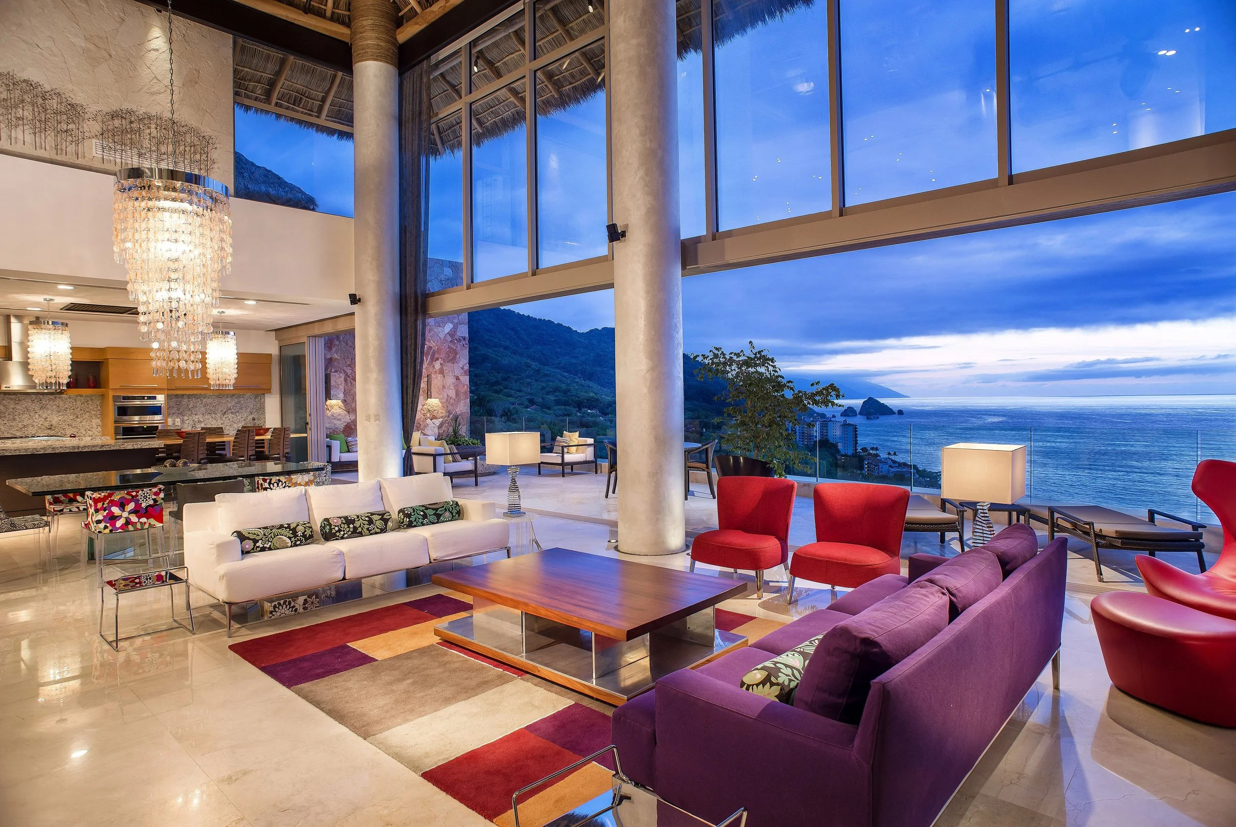 Interior of a modern living room with large windows overlooking the ocean and mountains, furnished with colorful sofas, armchairs, a wooden coffee table, chandeliers, and lamps.
