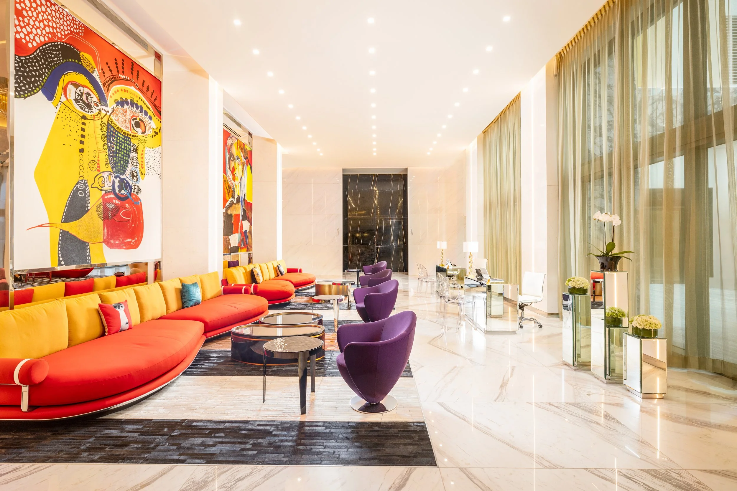 Elegant hotel lobby with colorful modern furniture, large abstract artwork on the wall, marble floors, and floor-to-ceiling curtains.