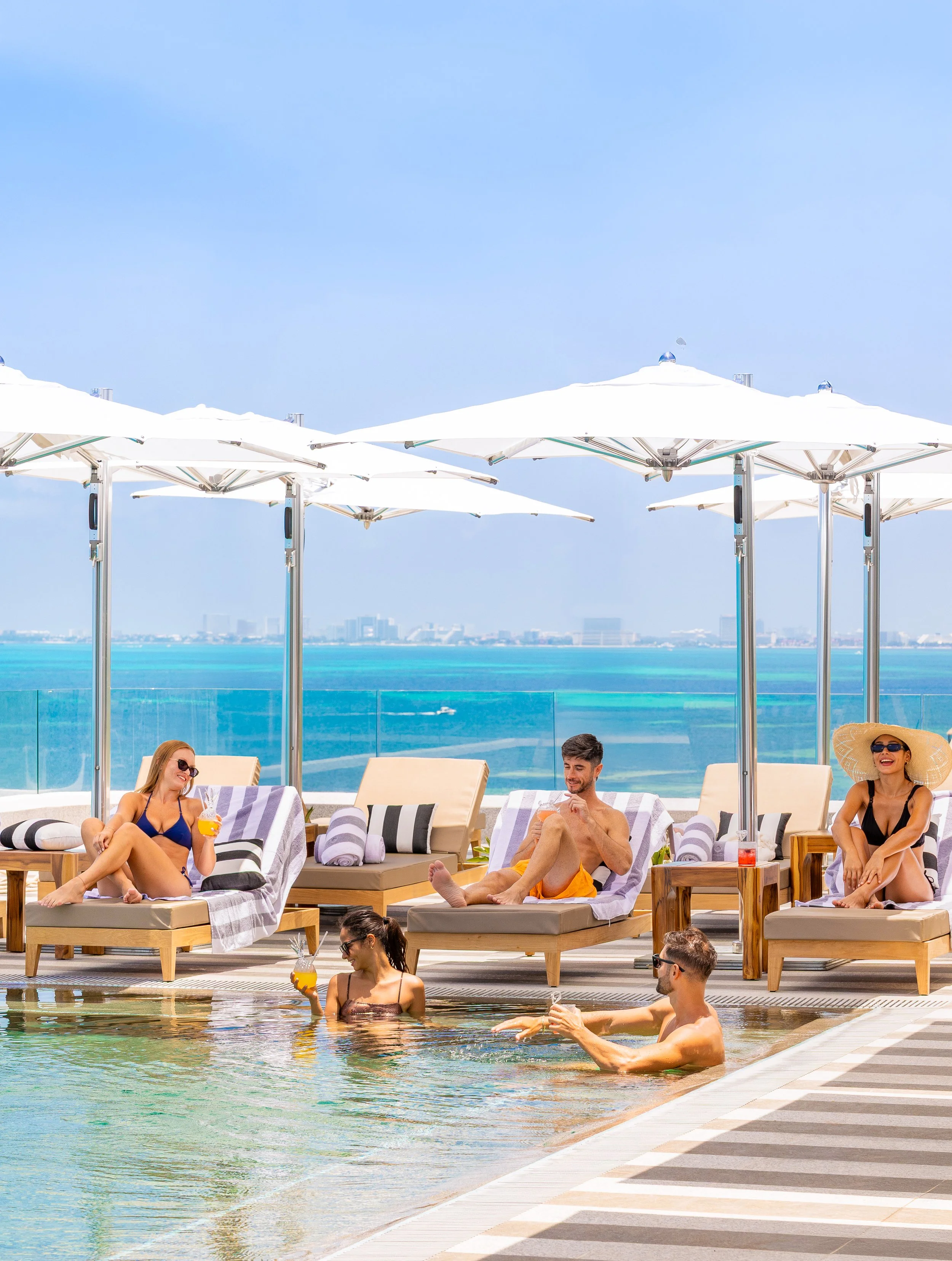 People relaxing and enjoying drinks by a pool on a rooftop with umbrellas, overlooking the ocean and city skyline.