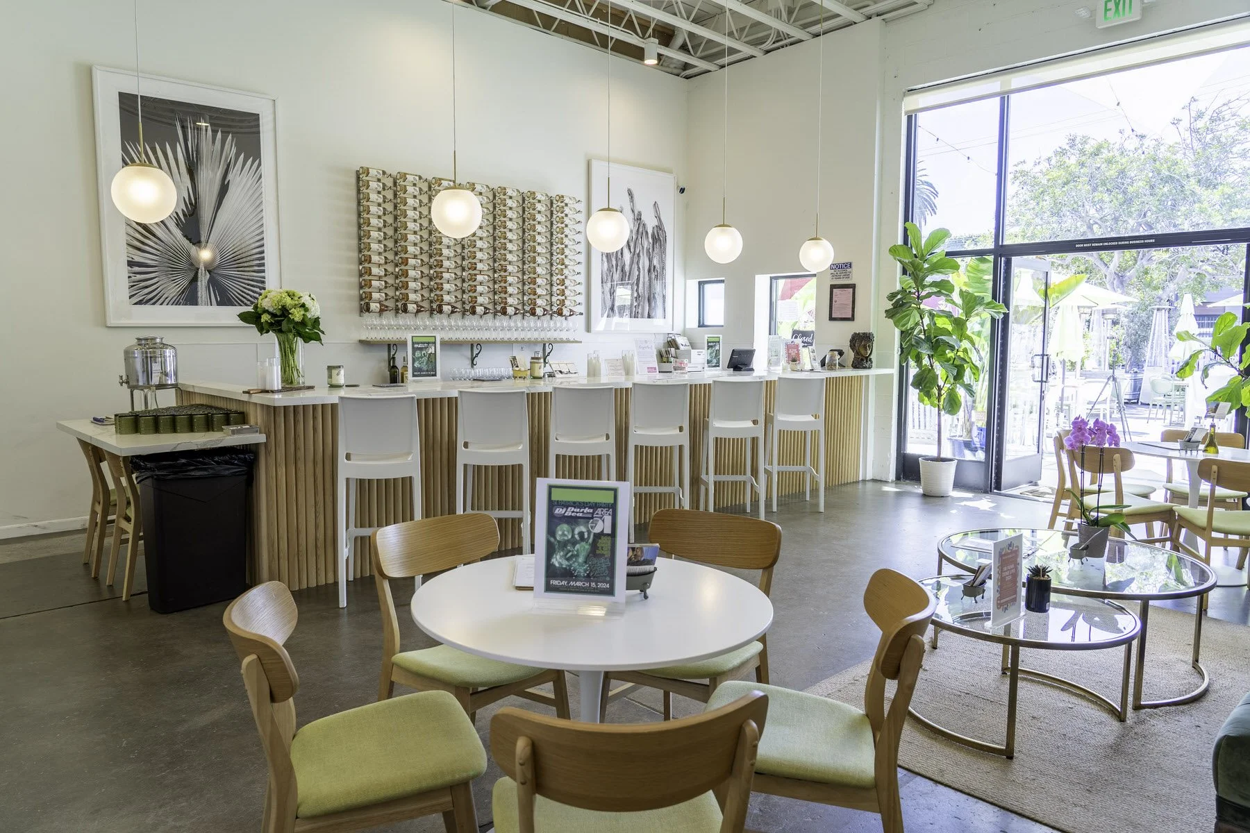 Bright, modern cafe interior with a white bar counter, barstools, round tables with chairs, large window with a view of outdoor seating, green plants, and decorative art on the walls.
