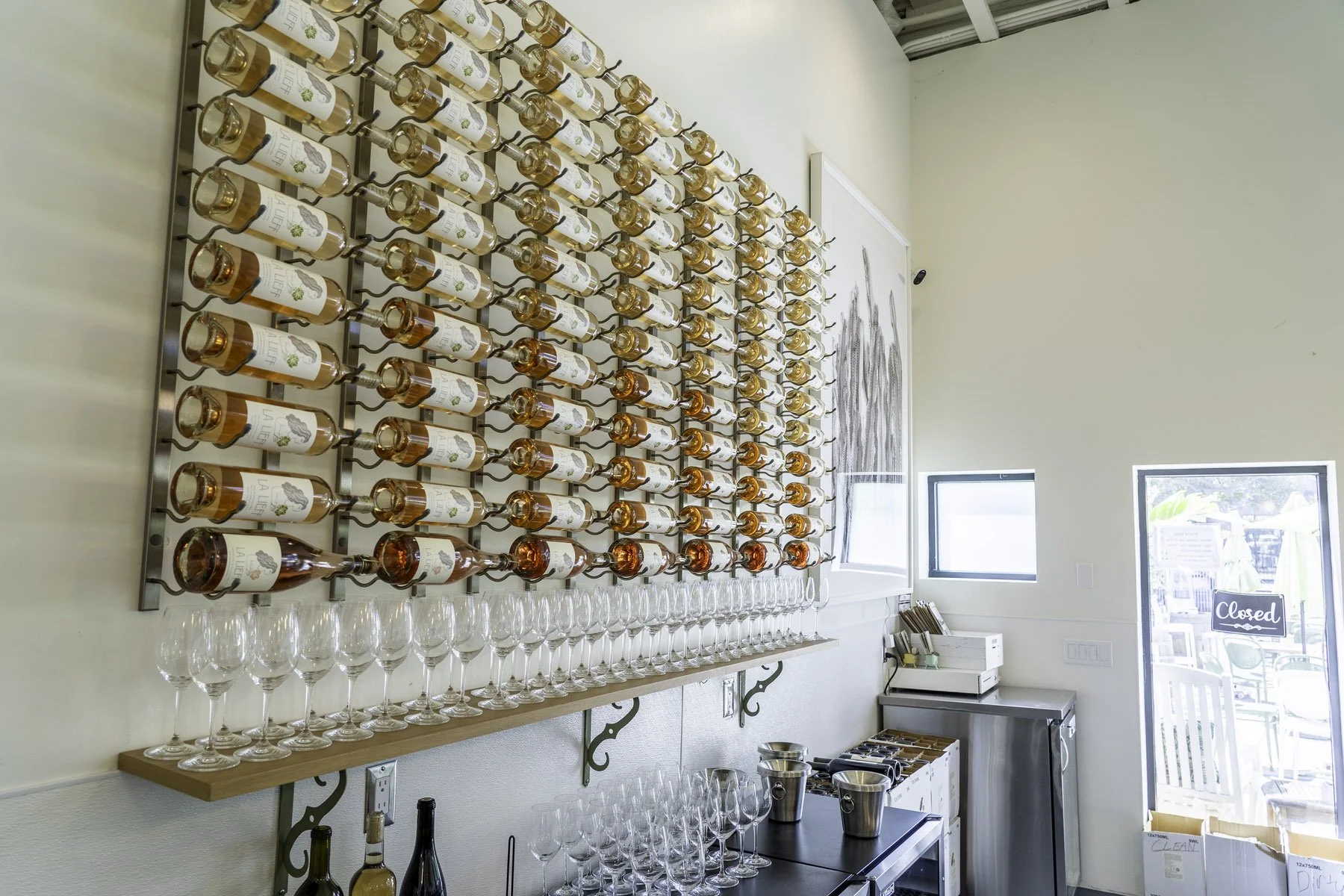 Wall-mounted display of multiple rows of wine bottles in a winery or tasting room, with empty wine glasses on a shelf below.