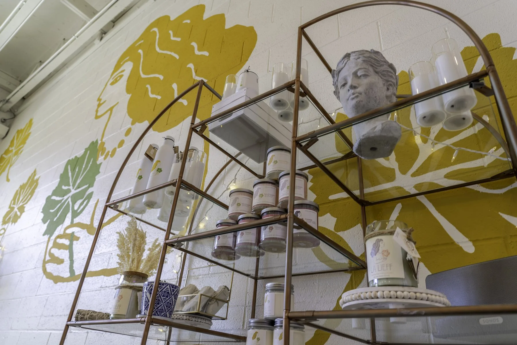 Interior of a store display shelf with candles, jars, and decorative items, against a wall with large yellow and green leaf artwork.
