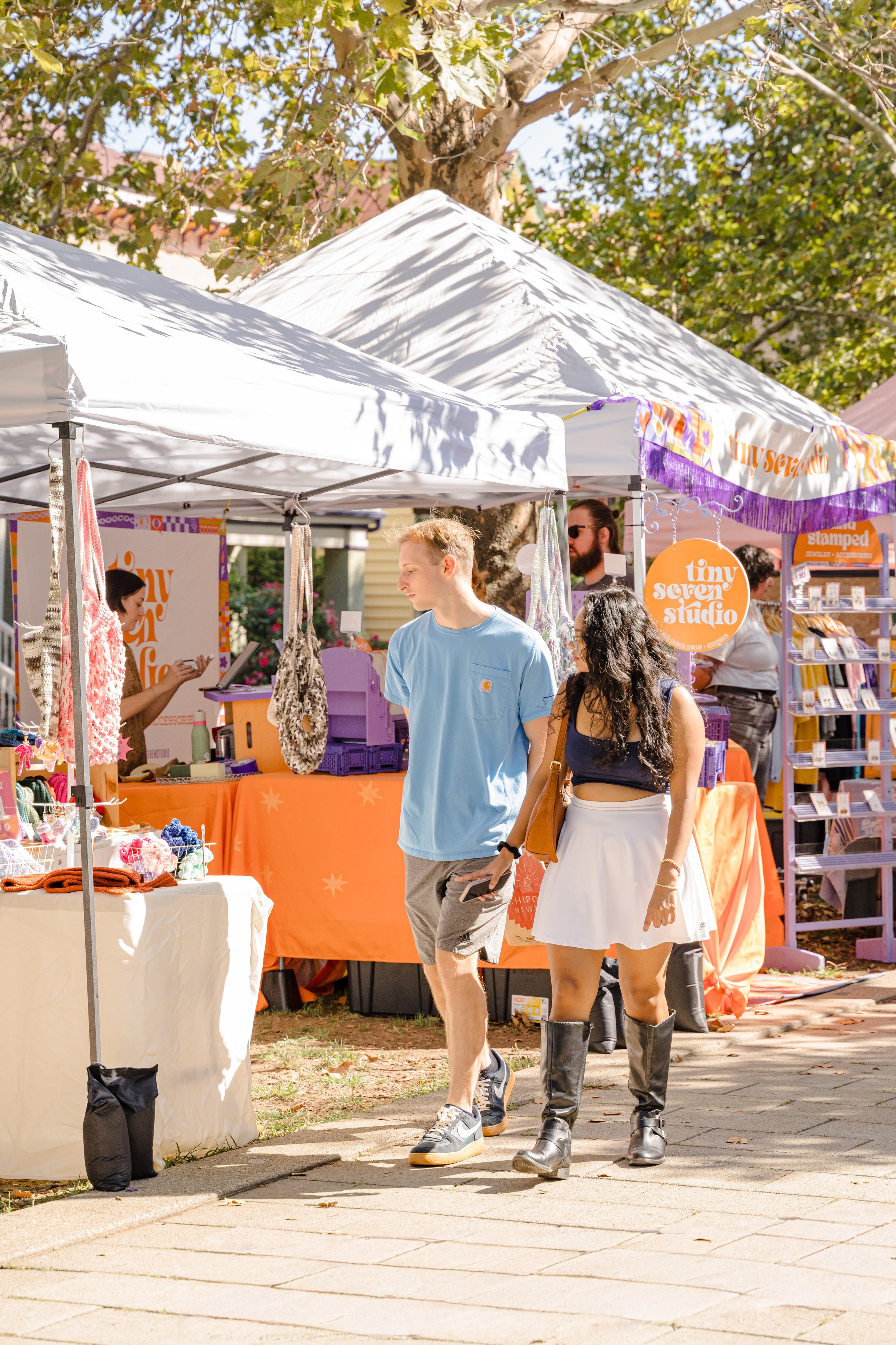 People shopping at an outdoor market with tents and vendor booths, surrounded by trees and sunlight.