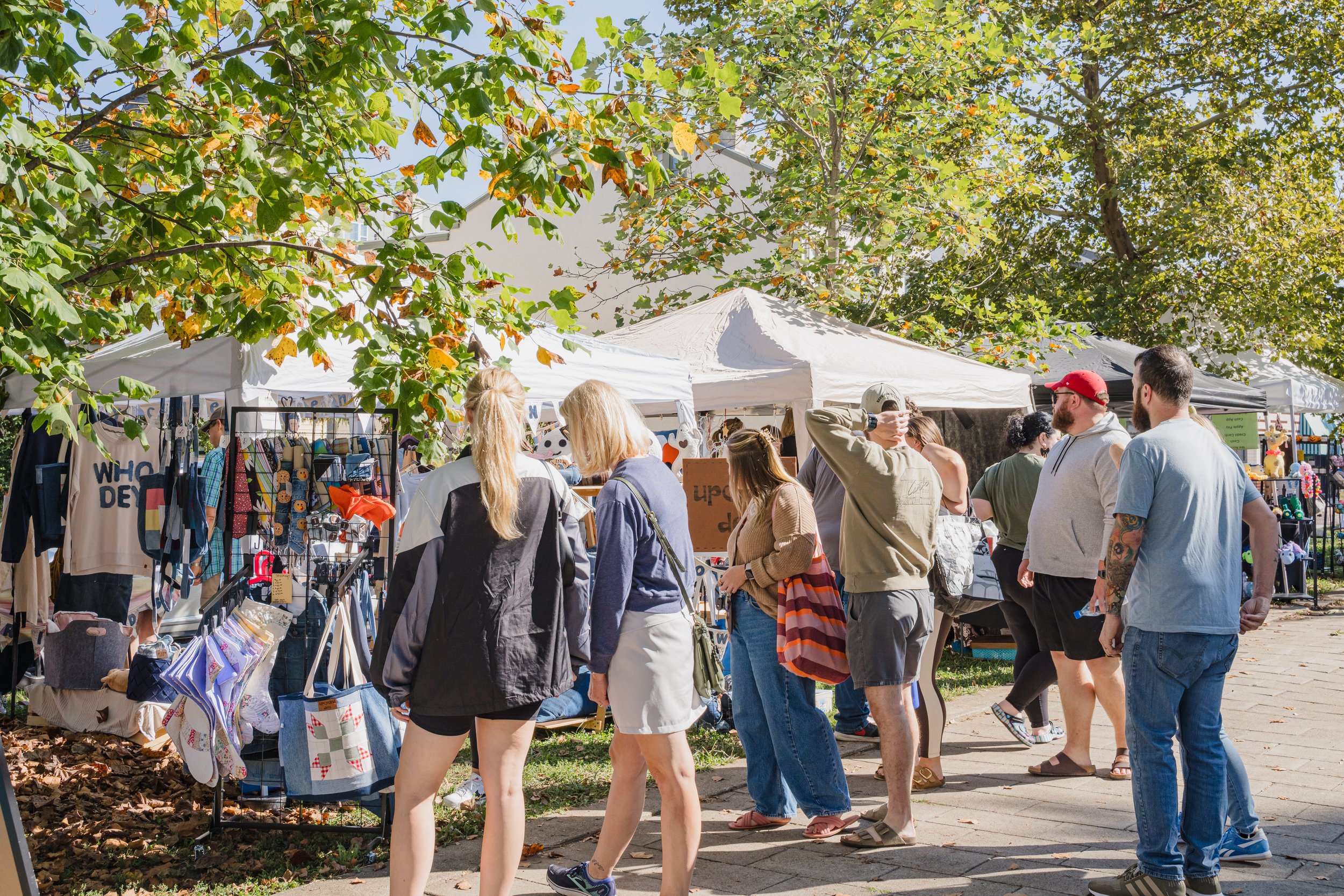 People shopping at outdoor market stalls on a sunny day with green trees in the background.
