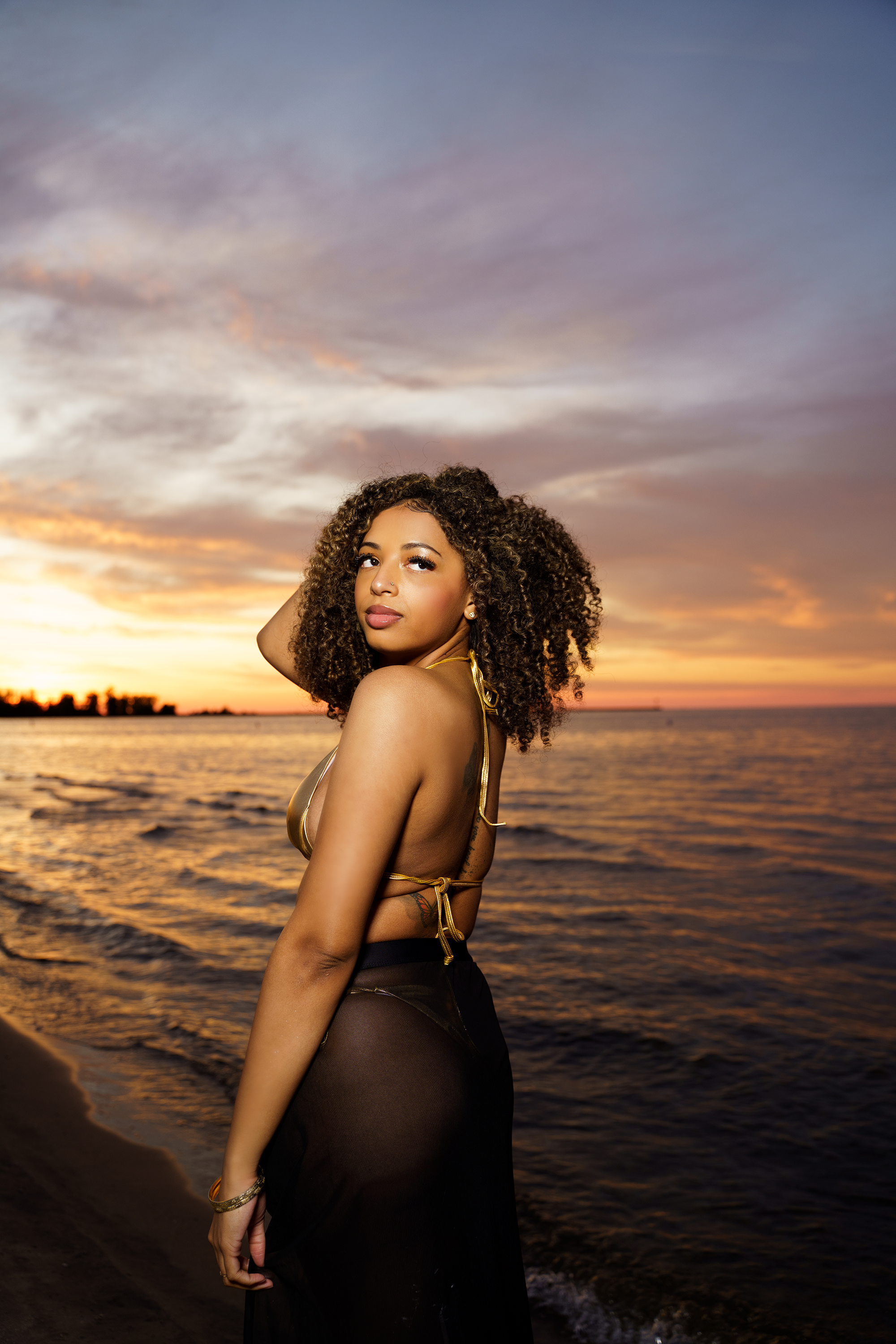 A woman with curly hair standing on a beach at sunset.