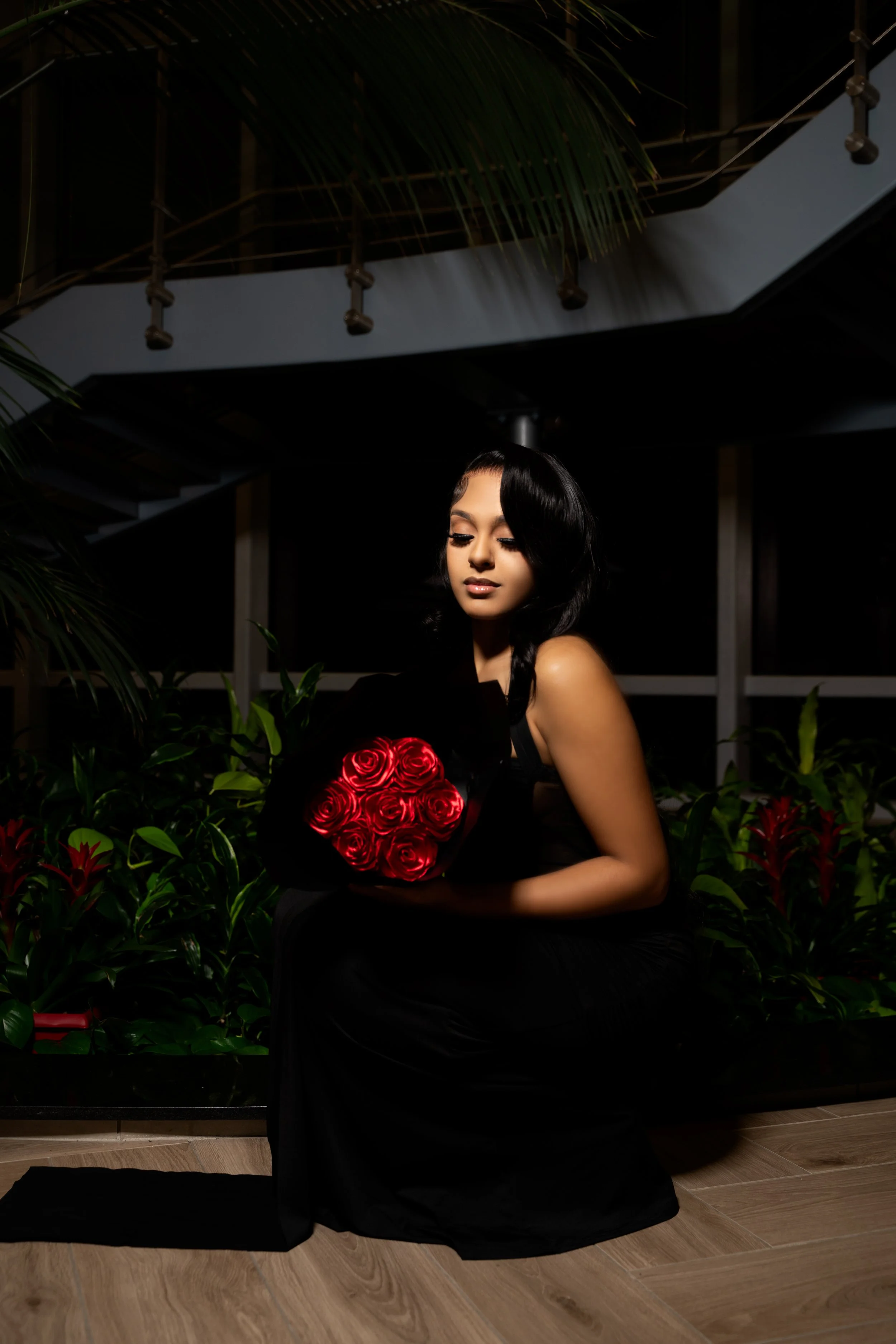 A woman in a black dress sitting on a wooden floor, holding a bouquet of red roses, with dark indoor surroundings and greenery in the background.