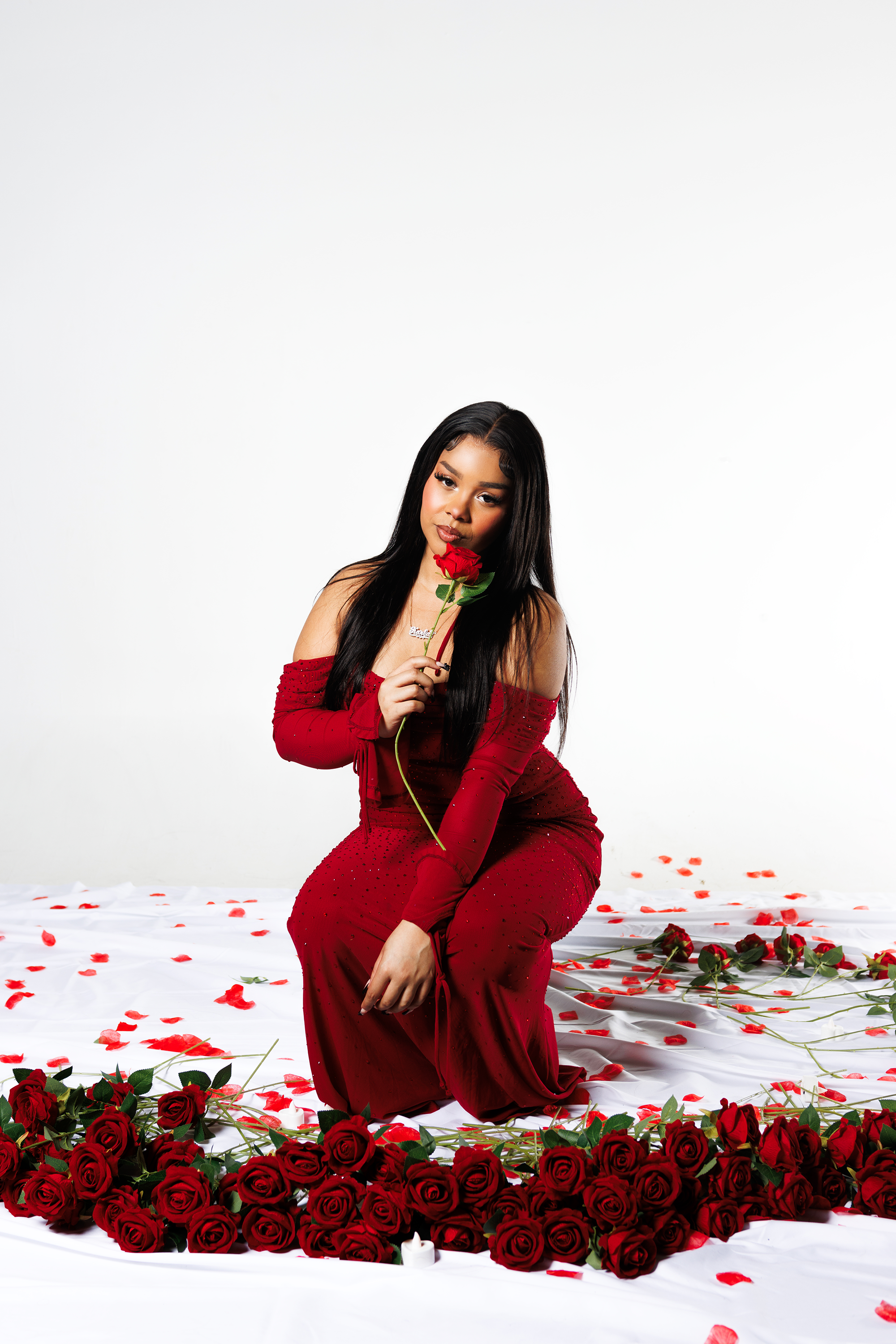 A woman in a red dress kneeling on a white sheet, surrounded by red roses and rose petals, holding a single stem of a rose near her face, with a plain white background.