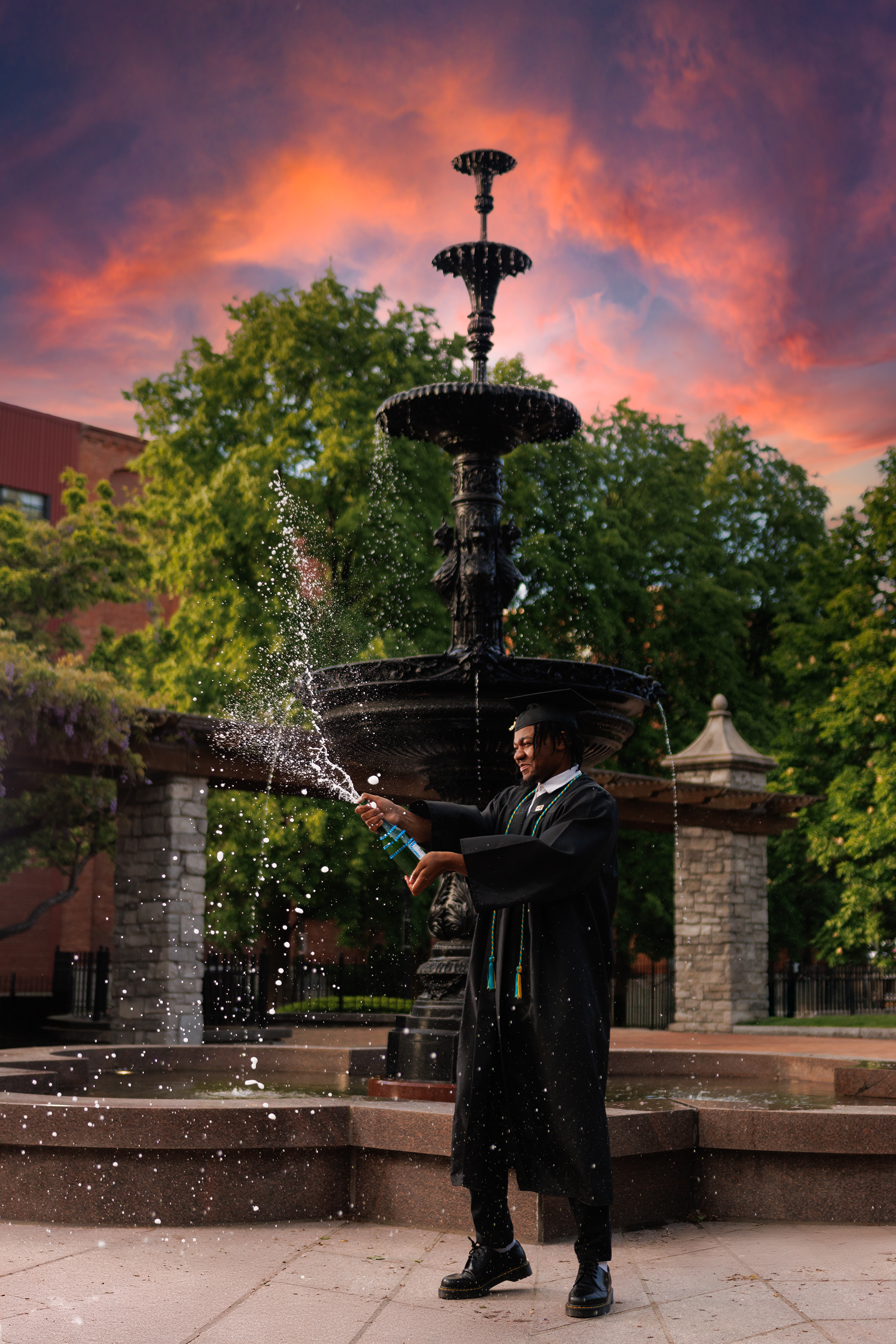 A graduate in a black cap and gown celebrating by splashing water from a bottle at a fountain during sunset.
