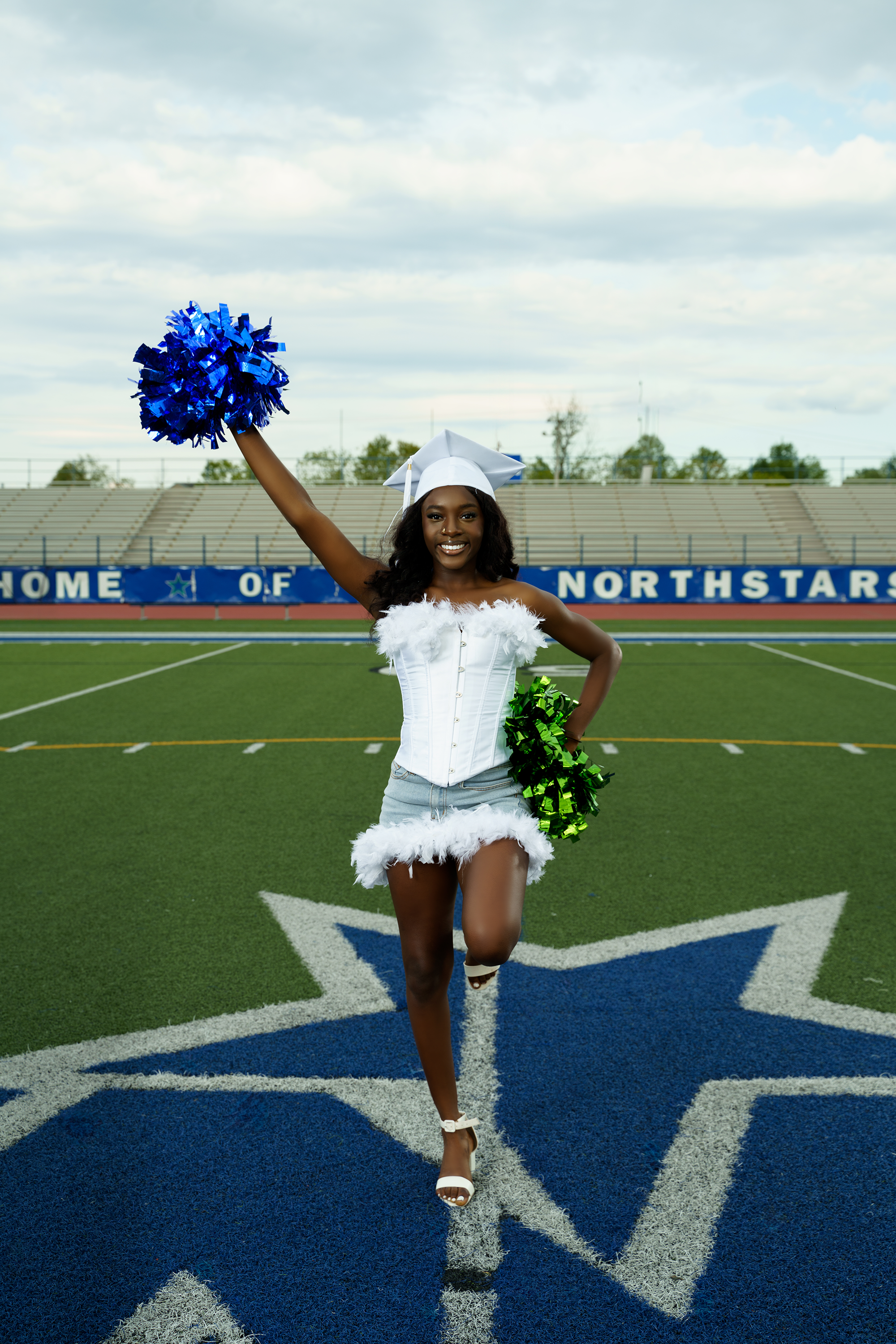 Young woman cheerleader on football field, wearing a white graduation cap and outfit with feather trim, holding blue and green pom-poms, standing on logo with star emblem, background stadium seats, and sky.