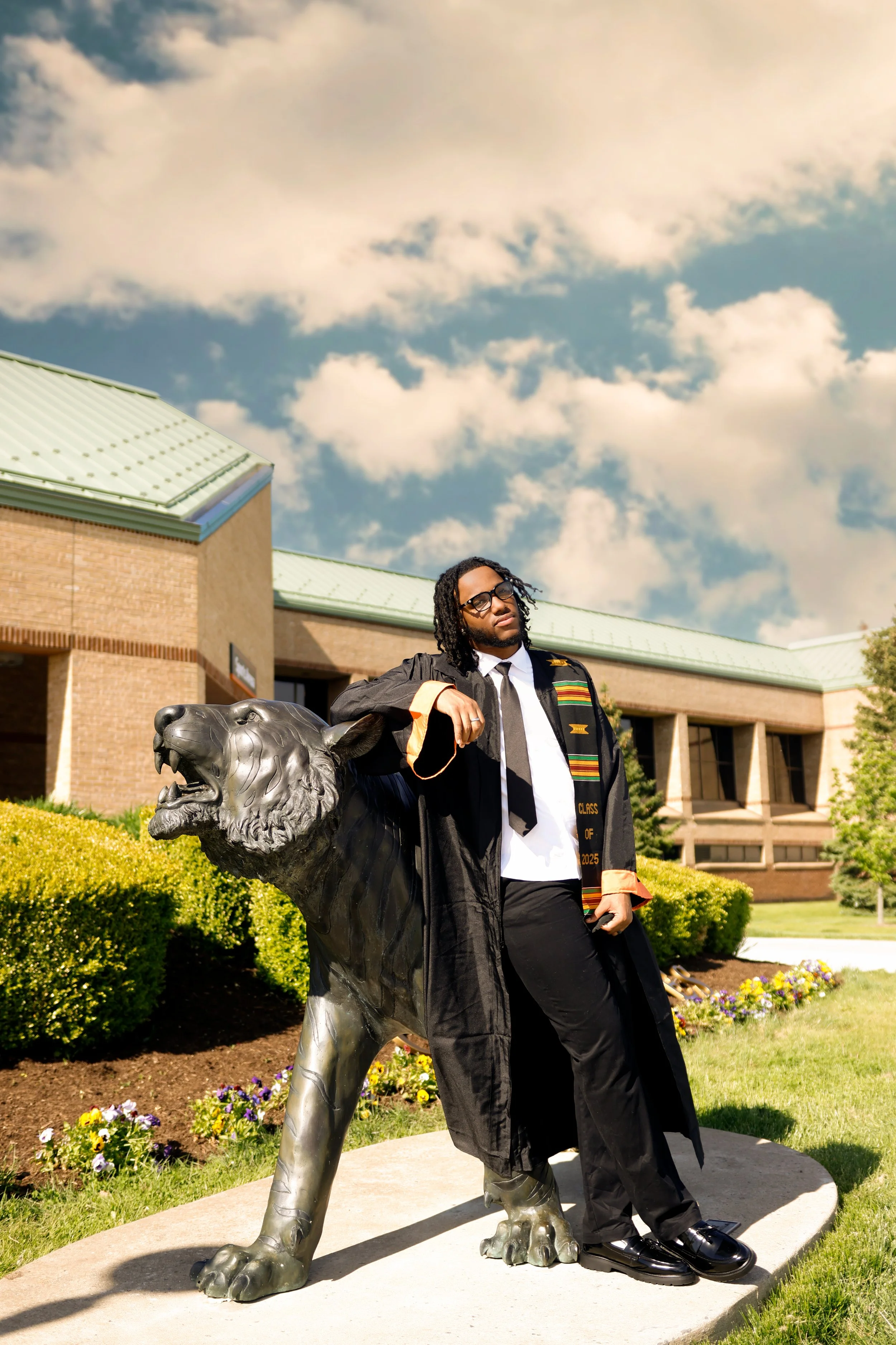 A man in graduation attire standing next to a large lion statue outside a school building with a green roof and blue sky with clouds in the background.