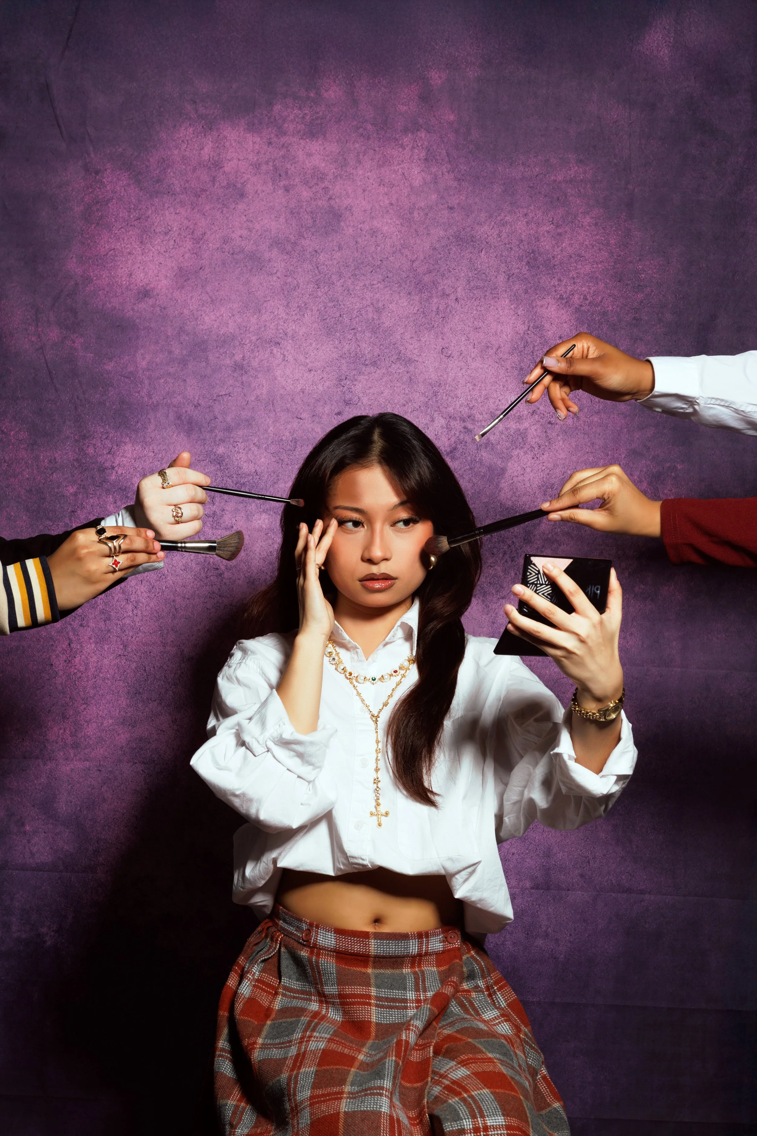 Woman wearing a white shirt with jewelry holds a mirror while makeup brushes are held near her face by multiple hands, against a purple textured background.