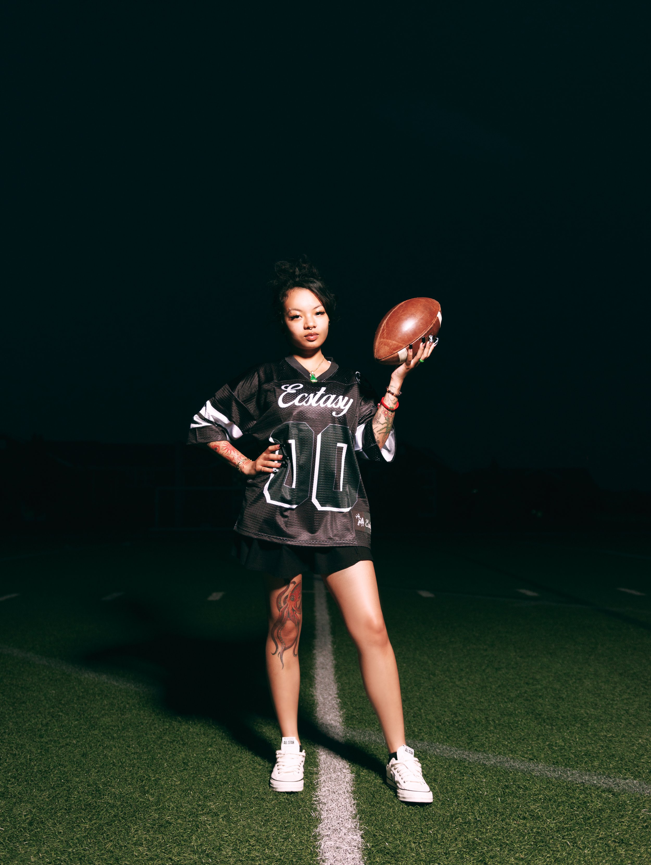 Young woman standing on a football field at night, holding a football in her right hand, wearing a black sports jersey with 'Ectasy' written on it, white sneakers, and sporting tattoos.
