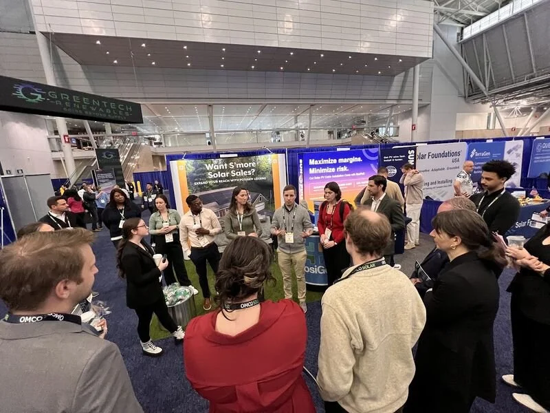 Group of people standing in a circle at a conference or expo, with booths and banners in the background promoting solar energy.